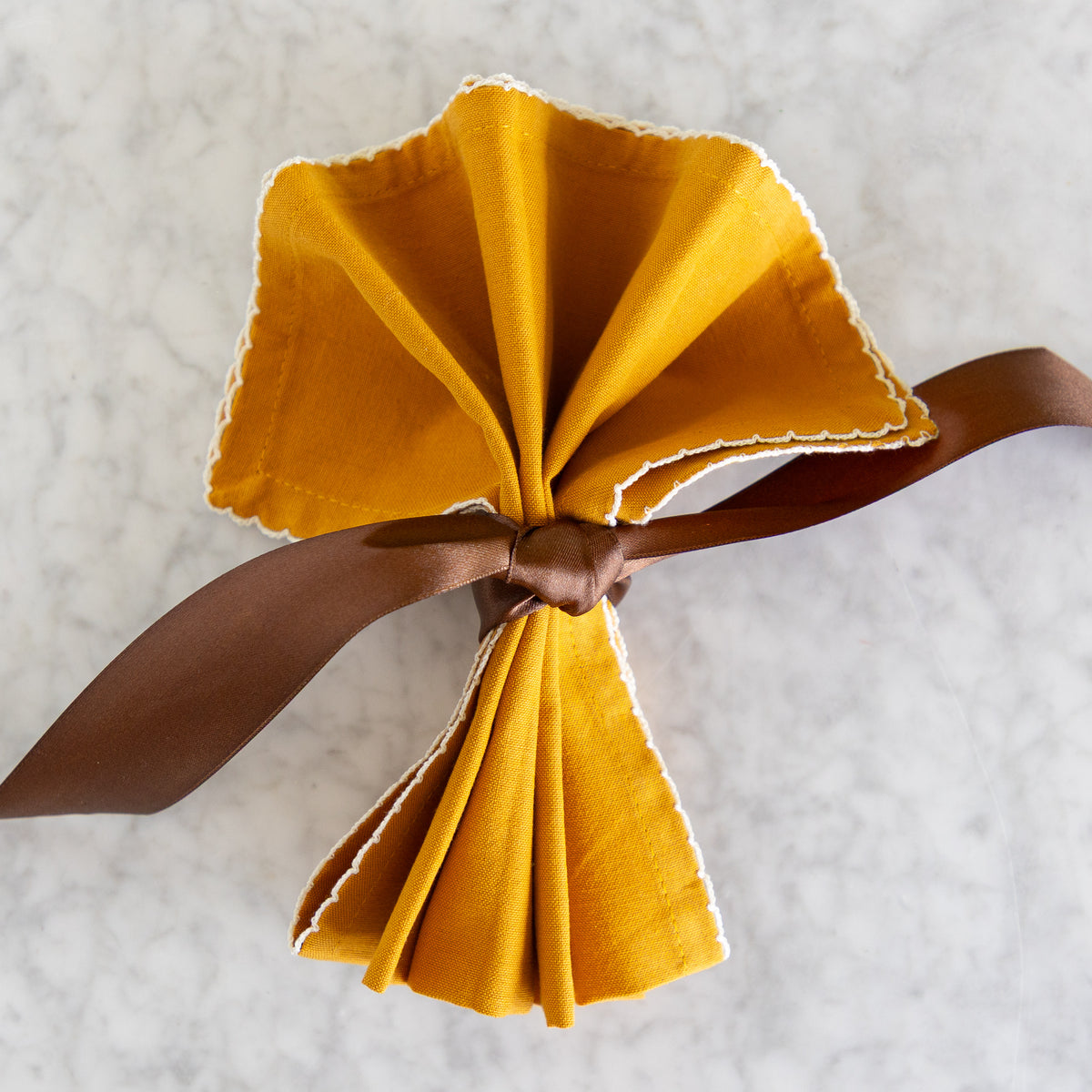 A folded mustard yellow fabric napkin with white scalloped edges, tied in a bow with a brown ribbon, placed on a white marble surface.