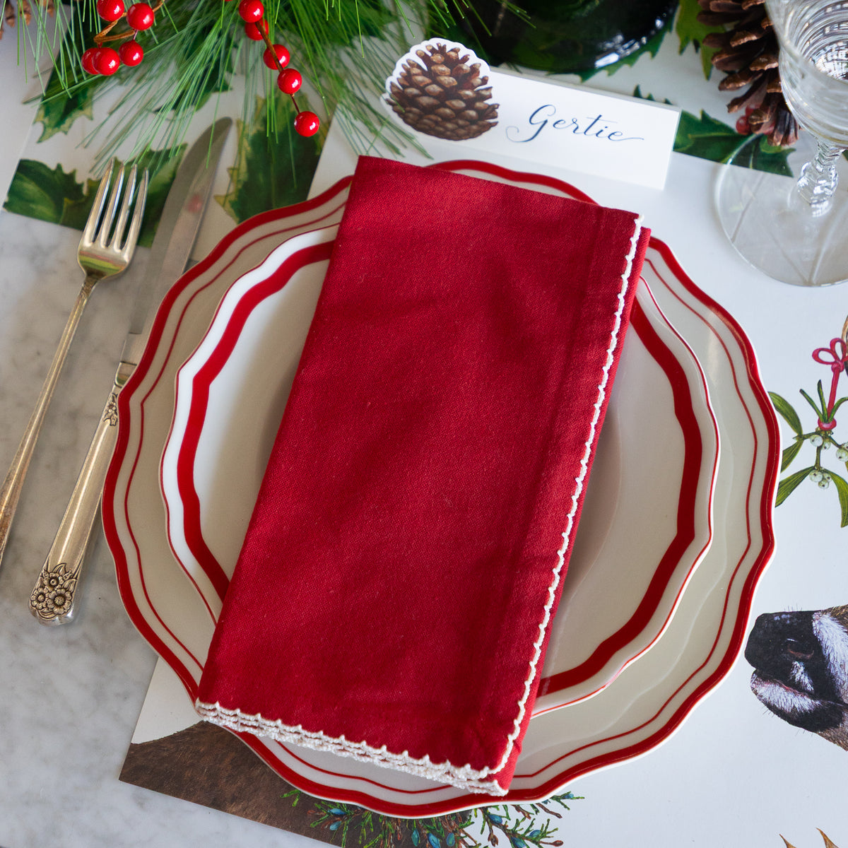 A festive holiday table setting featuring a deep red fabric napkin with white scalloped edges, layered over white plates with red borders, alongside gold-toned cutlery and holiday greenery.