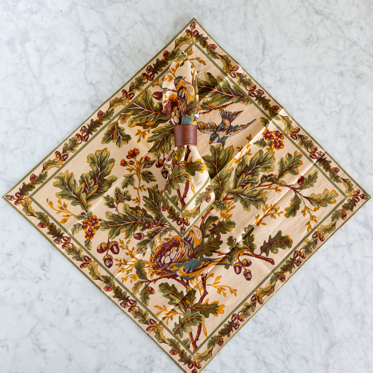 Cloth napkin with Fall colors and motif printed on it laying flat on a marble surface with another napkin held by a leather strap laying on top of it. 