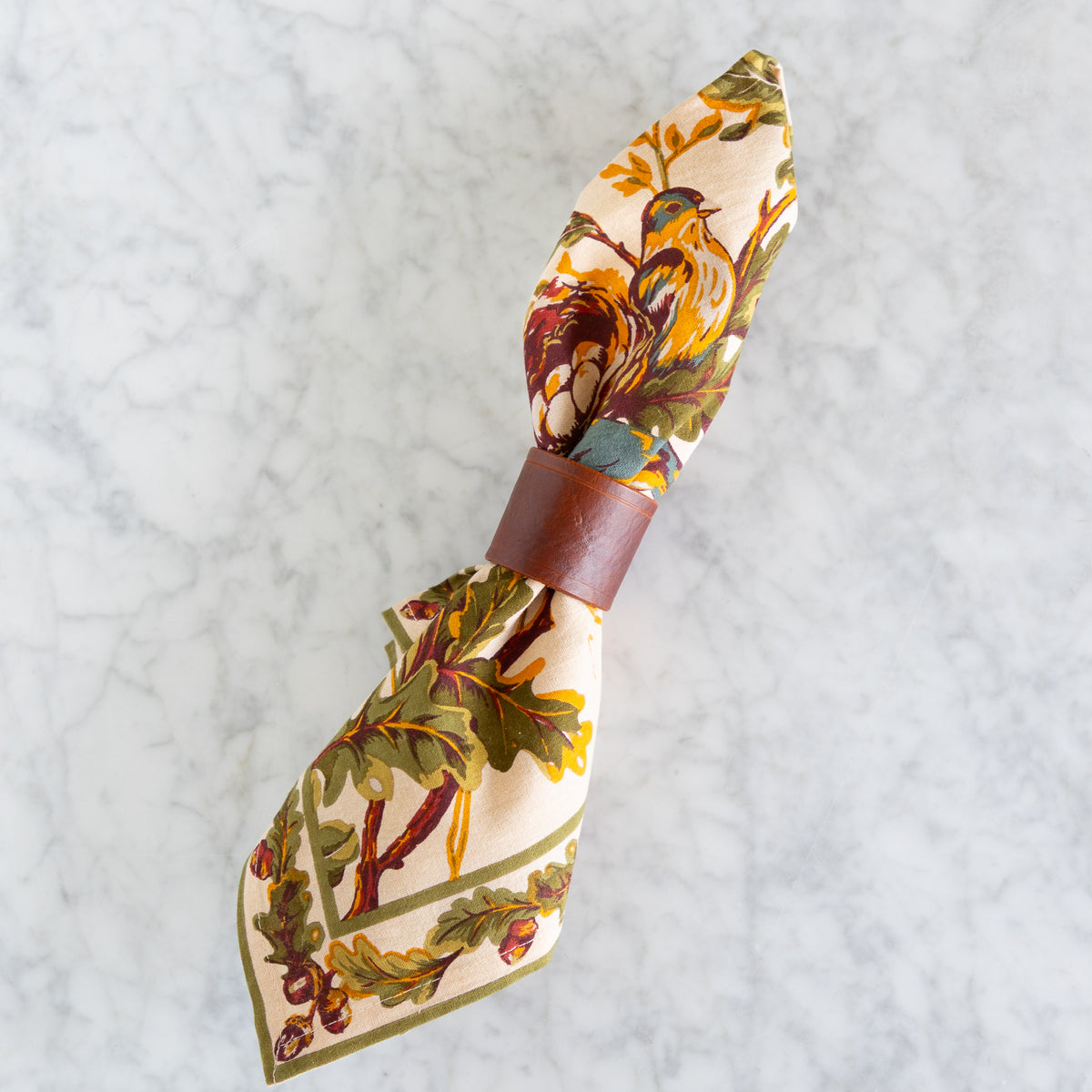 A fall-colored cotton napkin with an autumn leaf and acorn pattern, tied with a brown leather ring, displayed on a white marble surface.