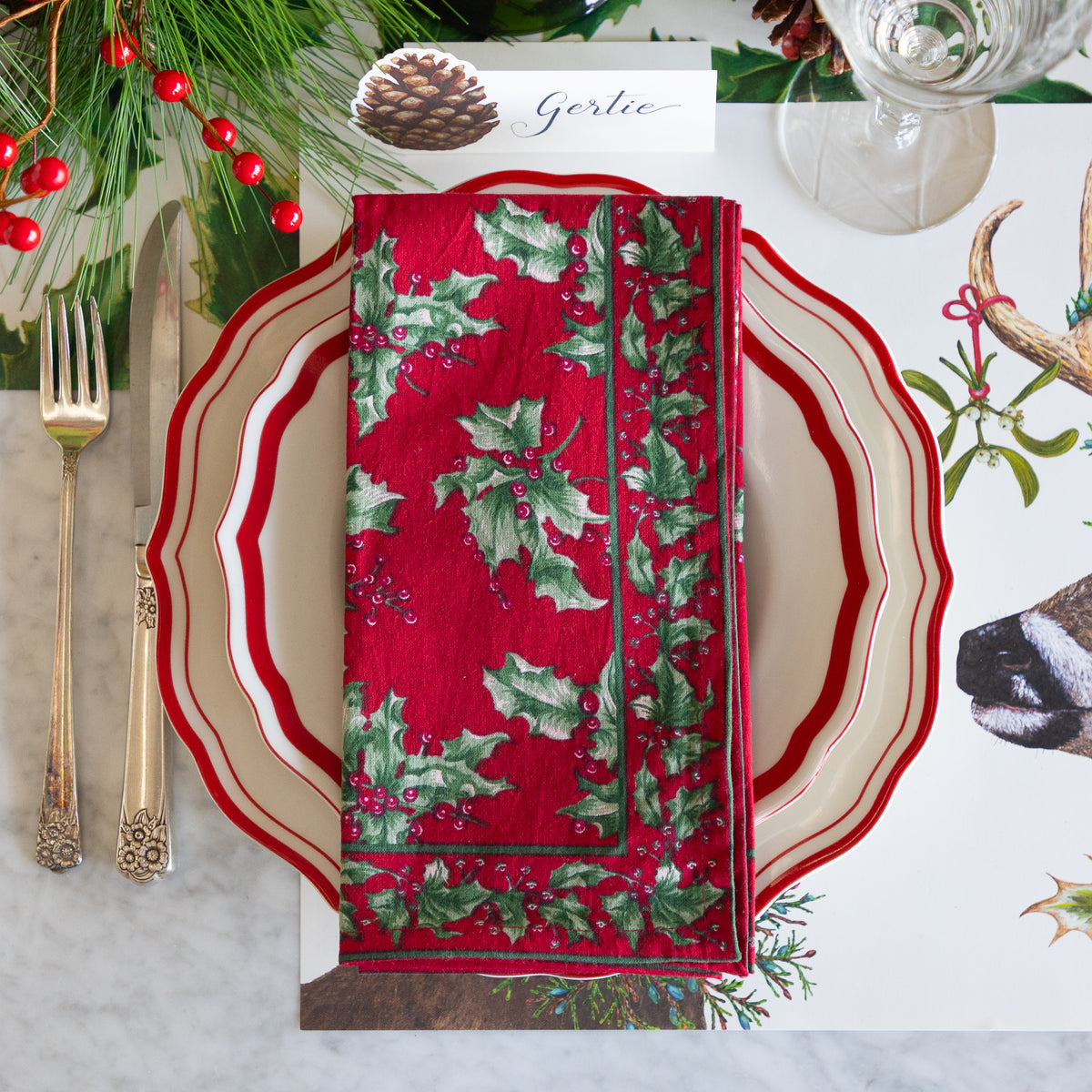 A festive holiday table setting featuring a red napkin with a green holly and berry pattern, layered on a cream and red scalloped charger plate. The setting includes gold-toned flatware, a glass, pinecones, evergreen branches, and a place card reading &