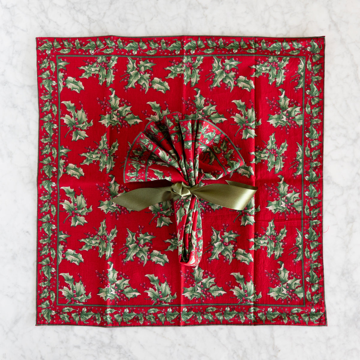 A square red cloth napkin with a green holly and berry border pattern, laying flat on a marble surface and tied with a shiny olive-green ribbon.