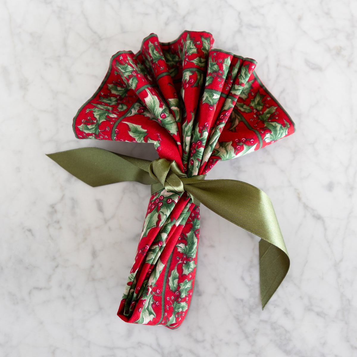 A folded red napkin with a green holly and berry pattern, tied with an olive green satin ribbon, displayed on a white marble surface.