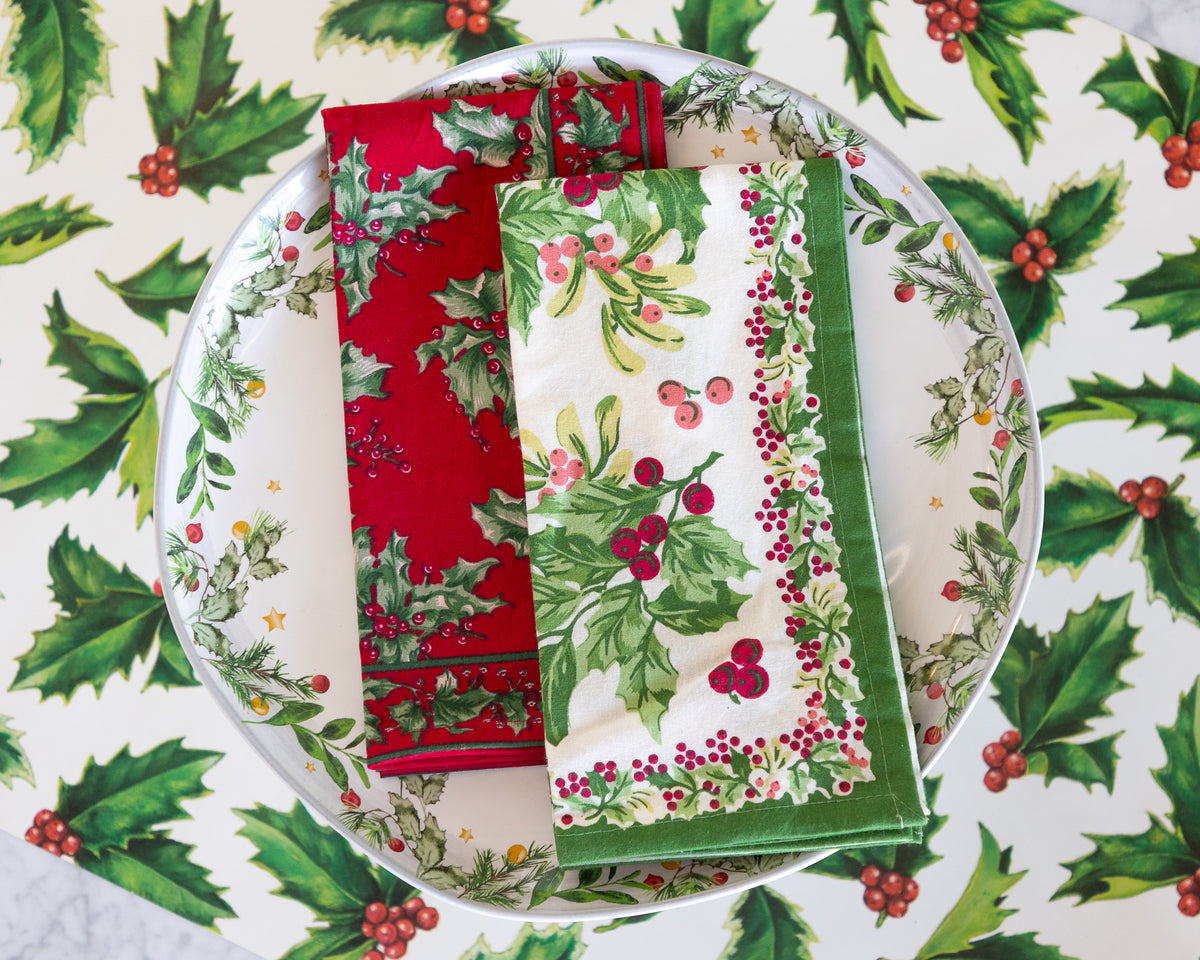 Two folded Christmas-themed fabric napkins with holly and berry patterns, displayed on a matching patterned plate.