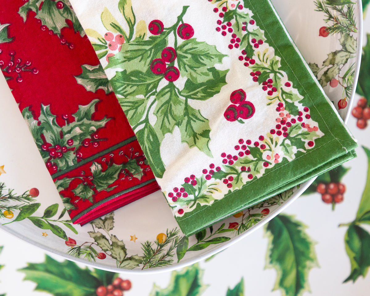 Two folded Christmas-themed fabric napkins with holly and berry patterns, displayed on a matching patterned plate.