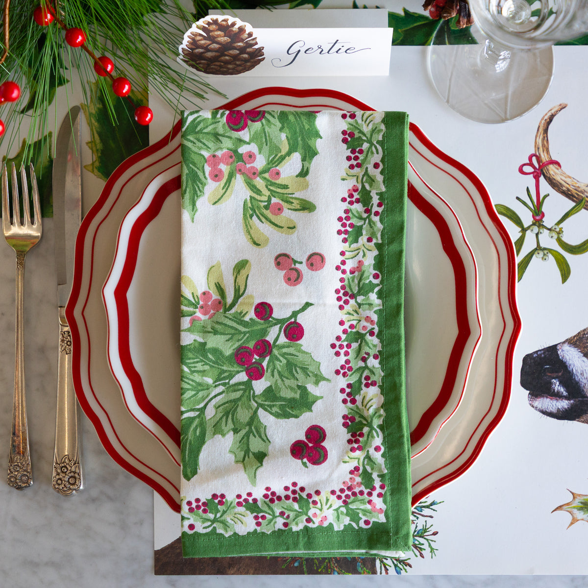 A festive Christmas table setting featuring a folded white napkin with a green holly and red berry pattern, layered over a red and white scalloped plate. The setting is styled with gold cutlery, a glass, pinecones, and holiday greenery on a white tablecloth printed with holly and a deer.
