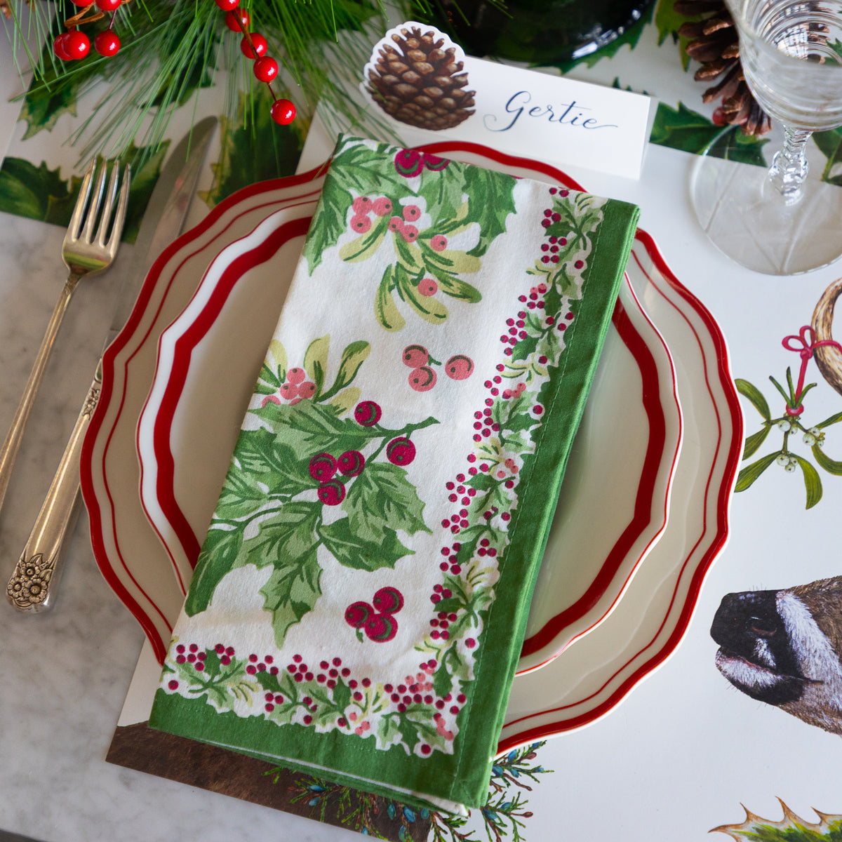 A festive Christmas table setting featuring a folded white napkin with a green holly and red berry pattern, layered over a red and white scalloped plate. The setting is styled with gold cutlery, a glass, pinecones, and holiday greenery on a white tablecloth printed with holly and a deer.