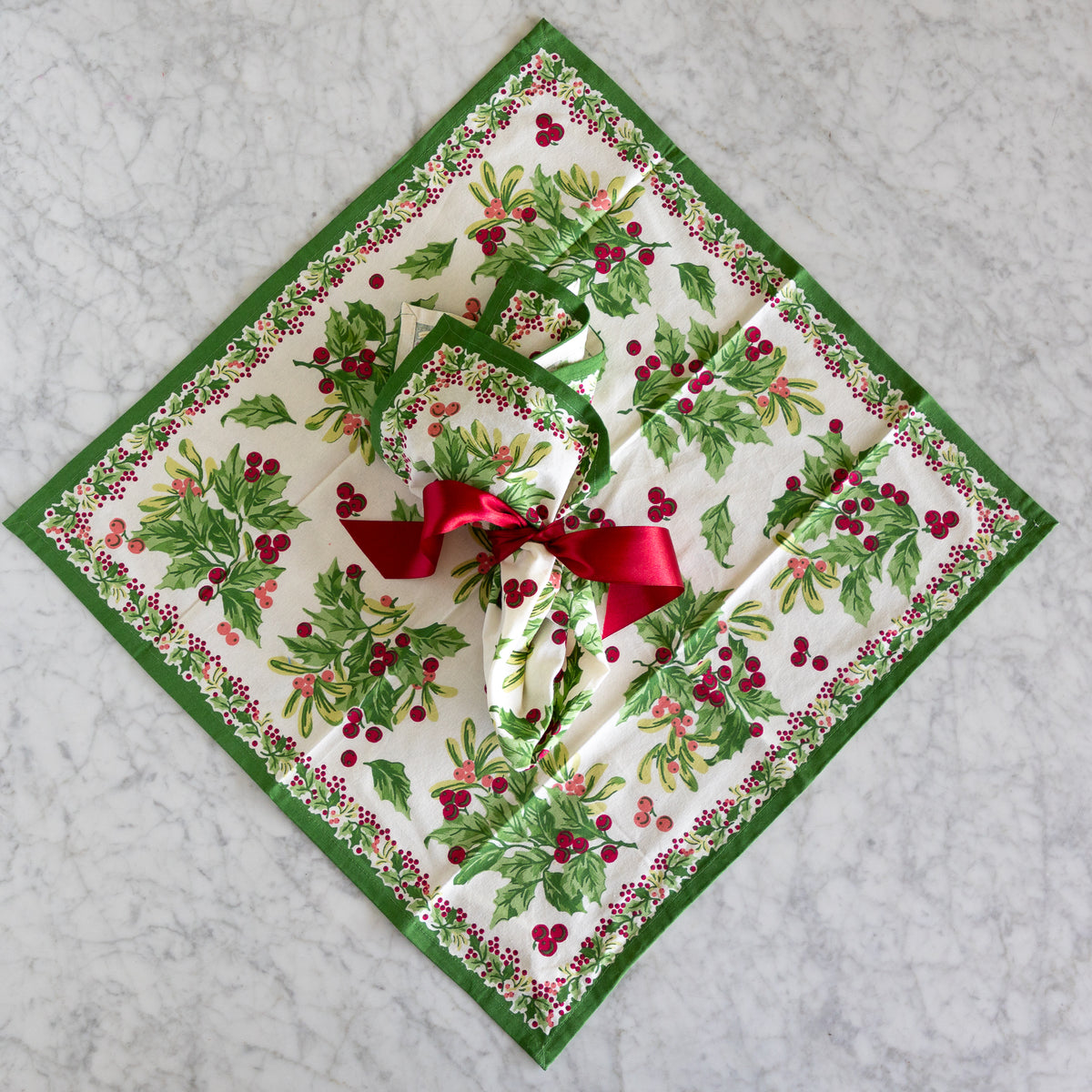 A square Christmas-themed tablecloth with a matching folded napkin and a red ribbon bow, displayed on a light gray marble surface. The fabric features a green border and a pattern of holly leaves and red berries on a white background.