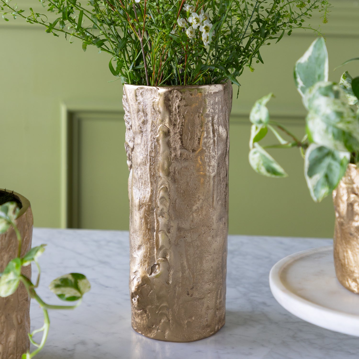 A tall Gold Birch Metal Vases with flowers on a marble surface with a green wall background.