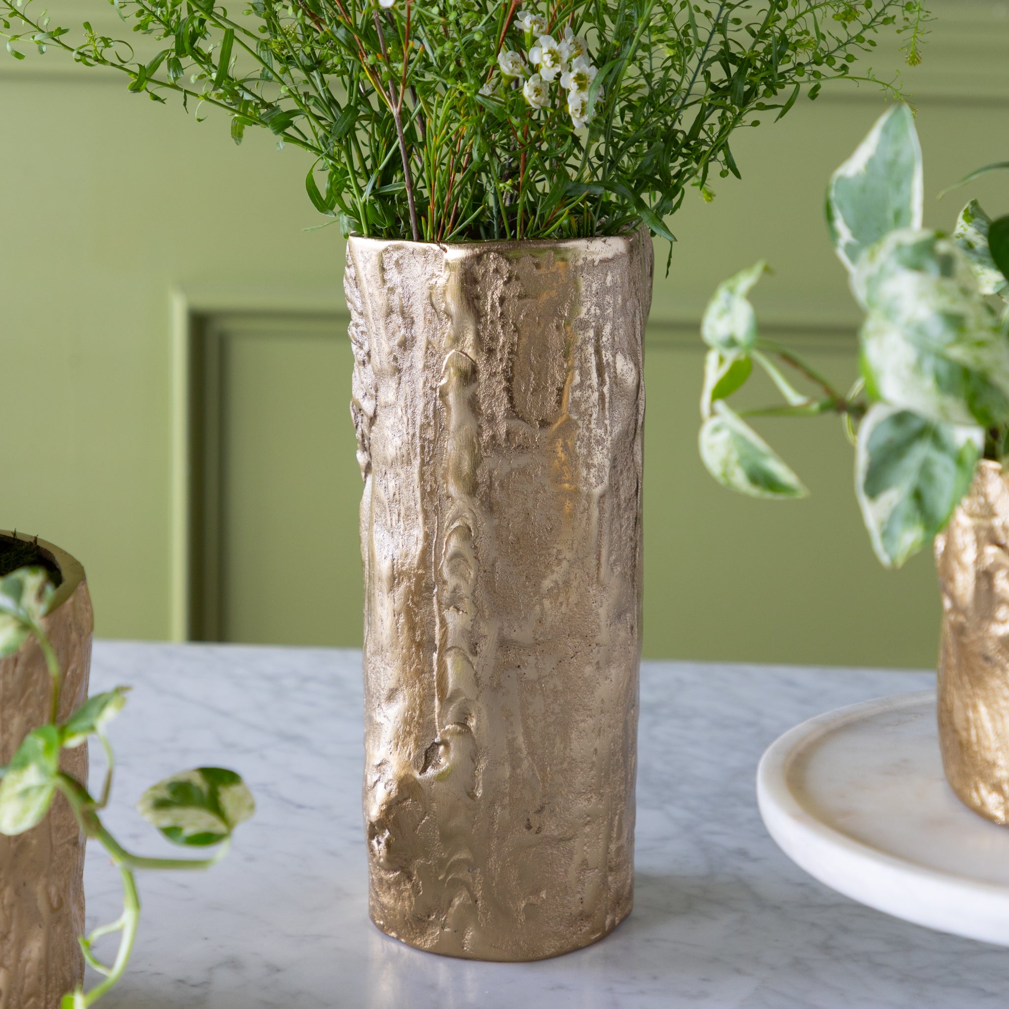 A tall Gold Birch Metal Vases with flowers on a marble surface with a green wall background.