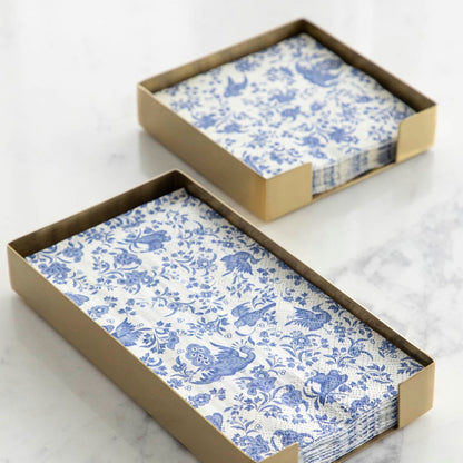 Two blue and white napkins elegantly placed in a handmade gold tray, accompanied by a Hester &amp; Cook brass napkin holder for added sophistication.