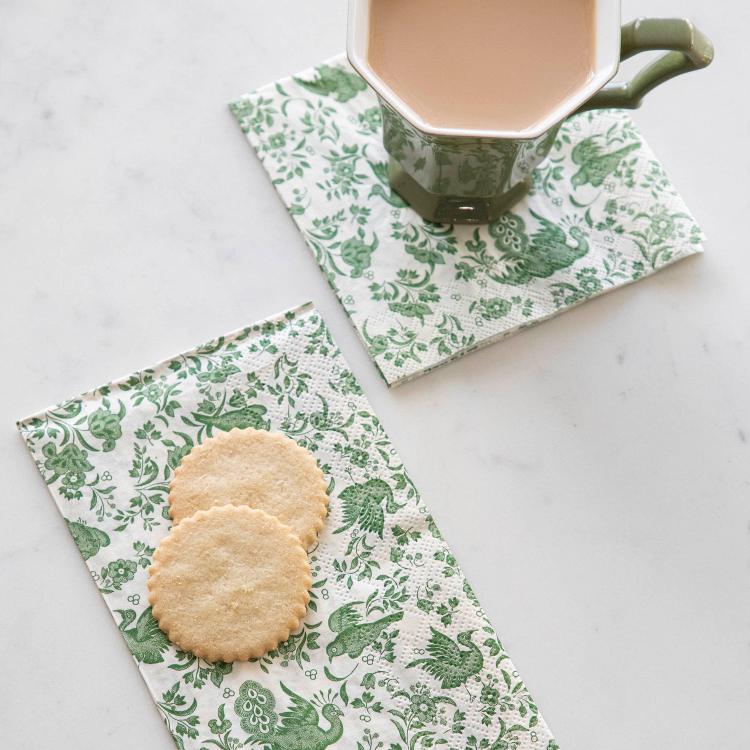 A cup of coffee and cookies on a table adorned with Hester &amp; Cook Green Regal Peacock napkins.