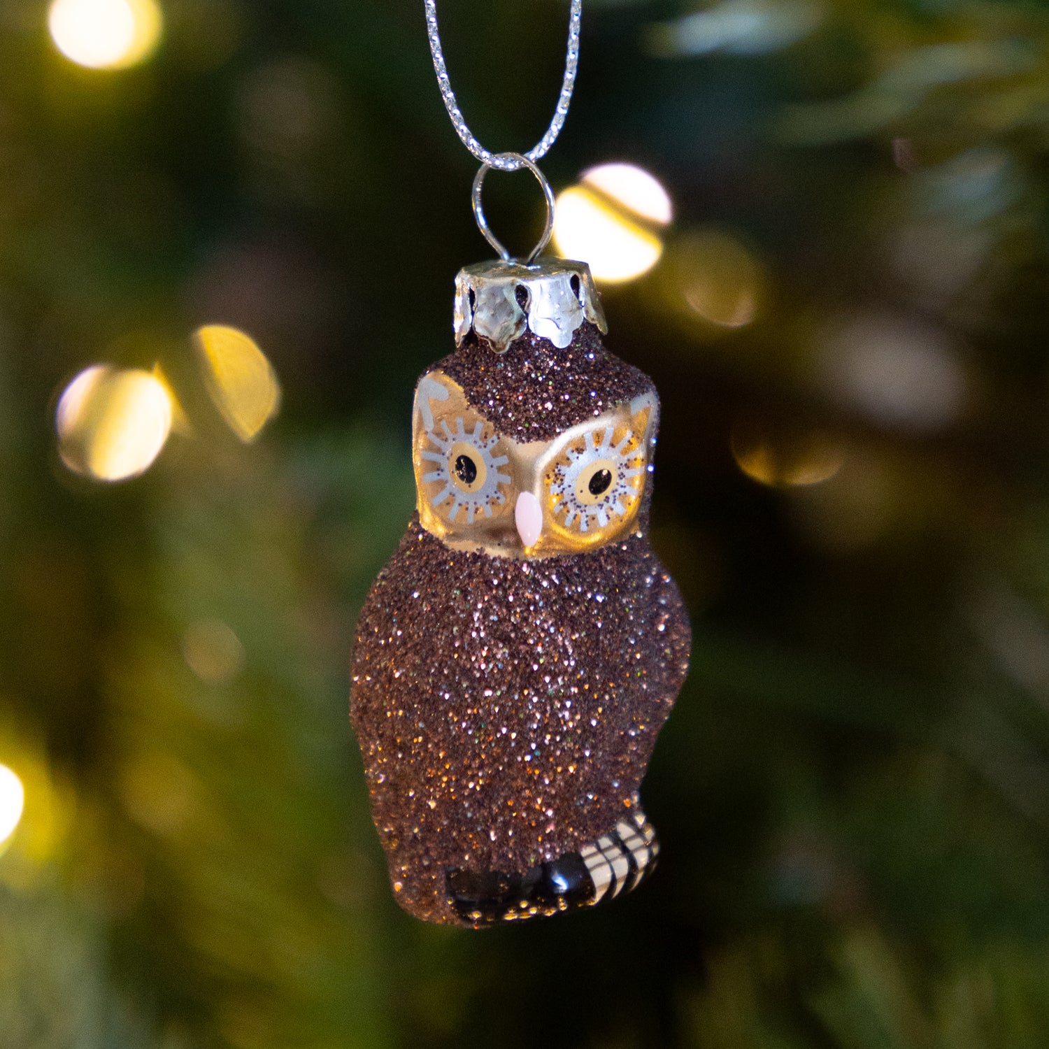 Sparkly brown owl ornament hanging on a tree with blurred lights in the background.