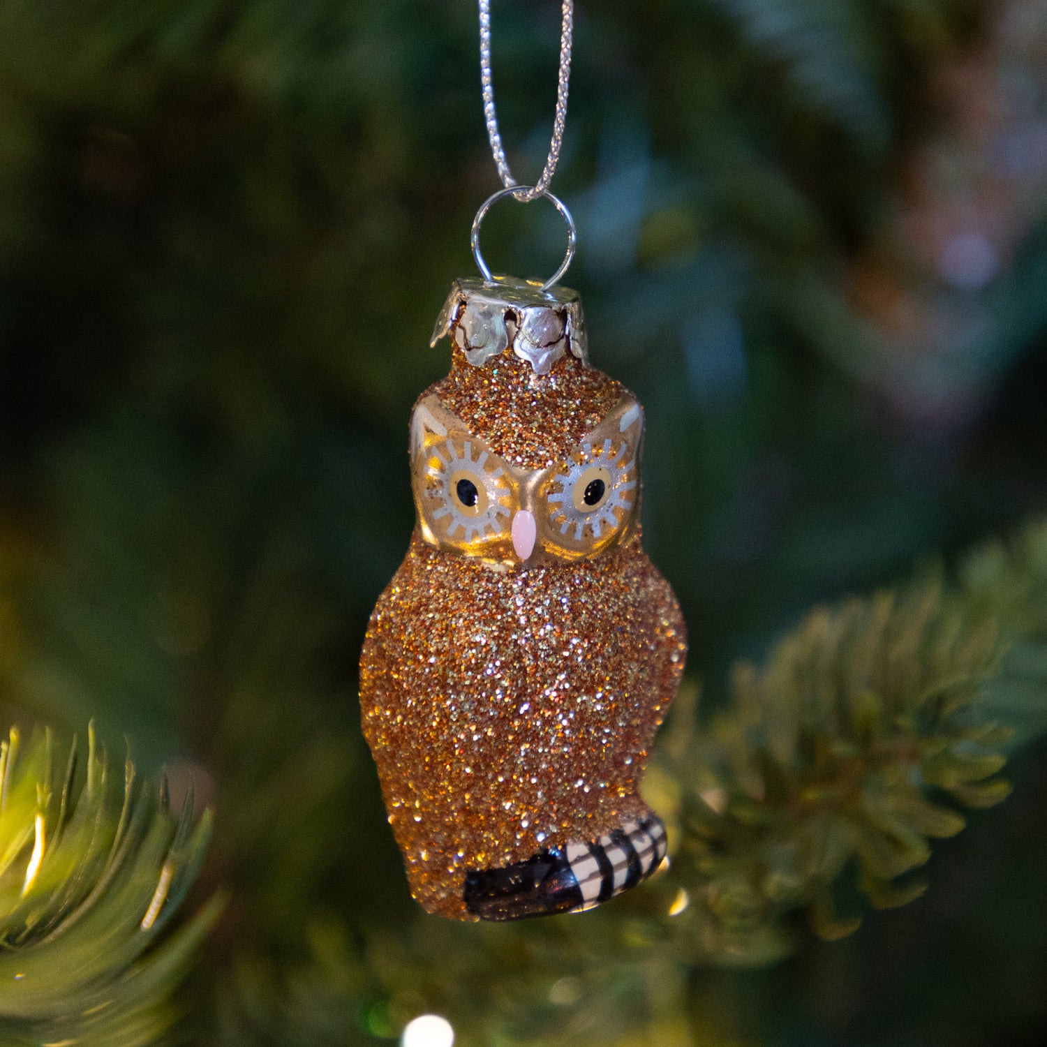 Sparkly copper owl ornament hanging on a tree with blurred lights in the background.