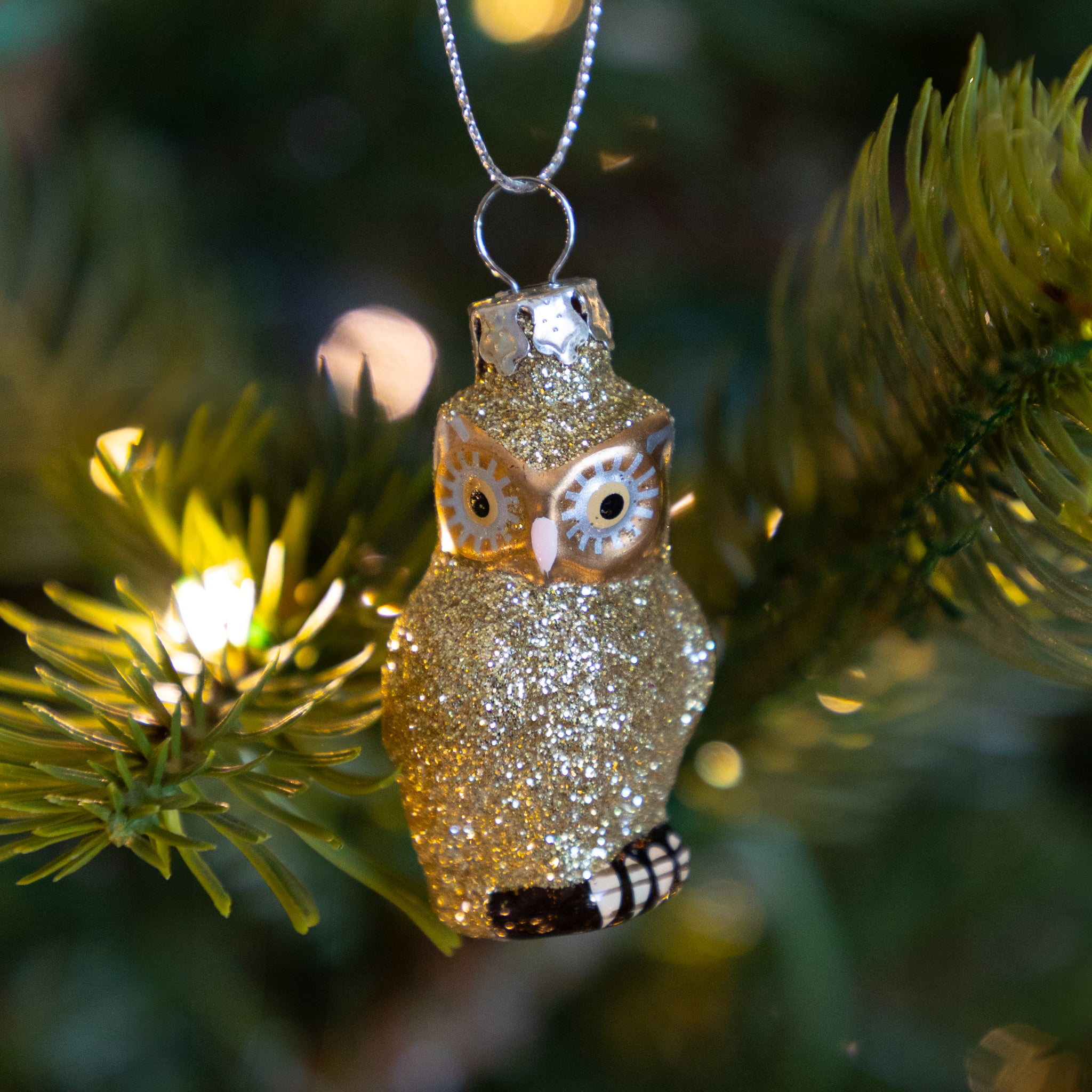Sparkly gold owl ornament hanging on a tree with blurred lights in the background.