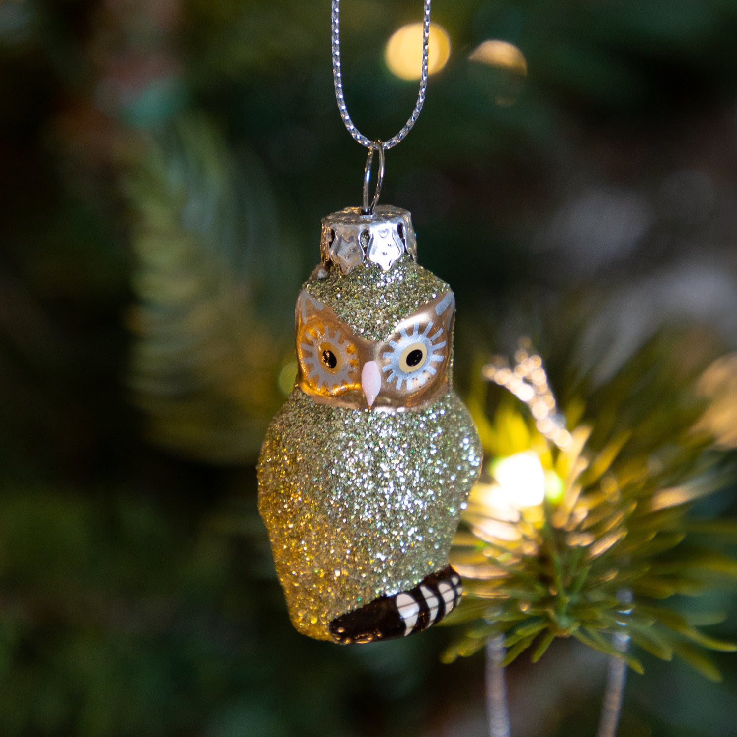 Sparkly green owl ornament hanging on a tree with blurred lights in the background.