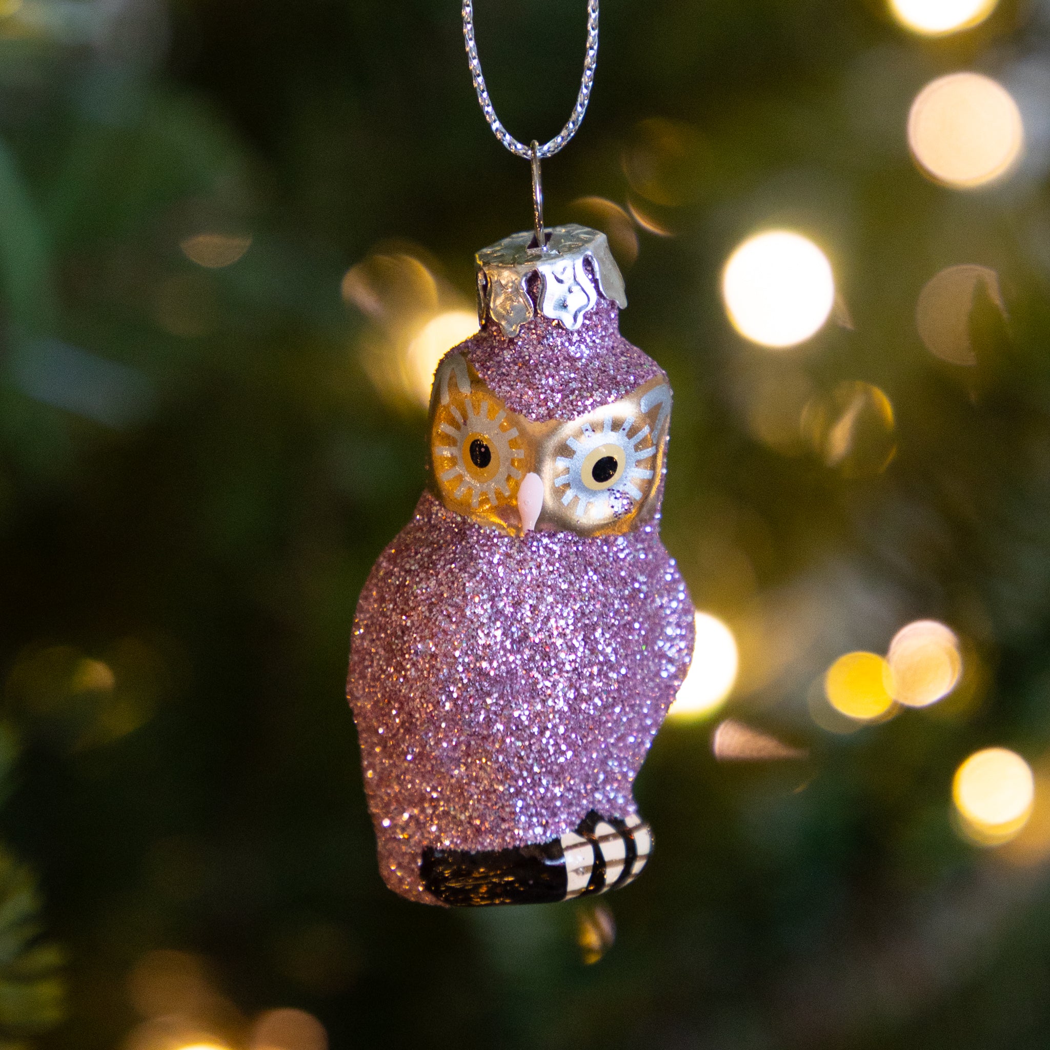 Sparkly pink owl ornament hanging on a tree with blurred lights in the background.