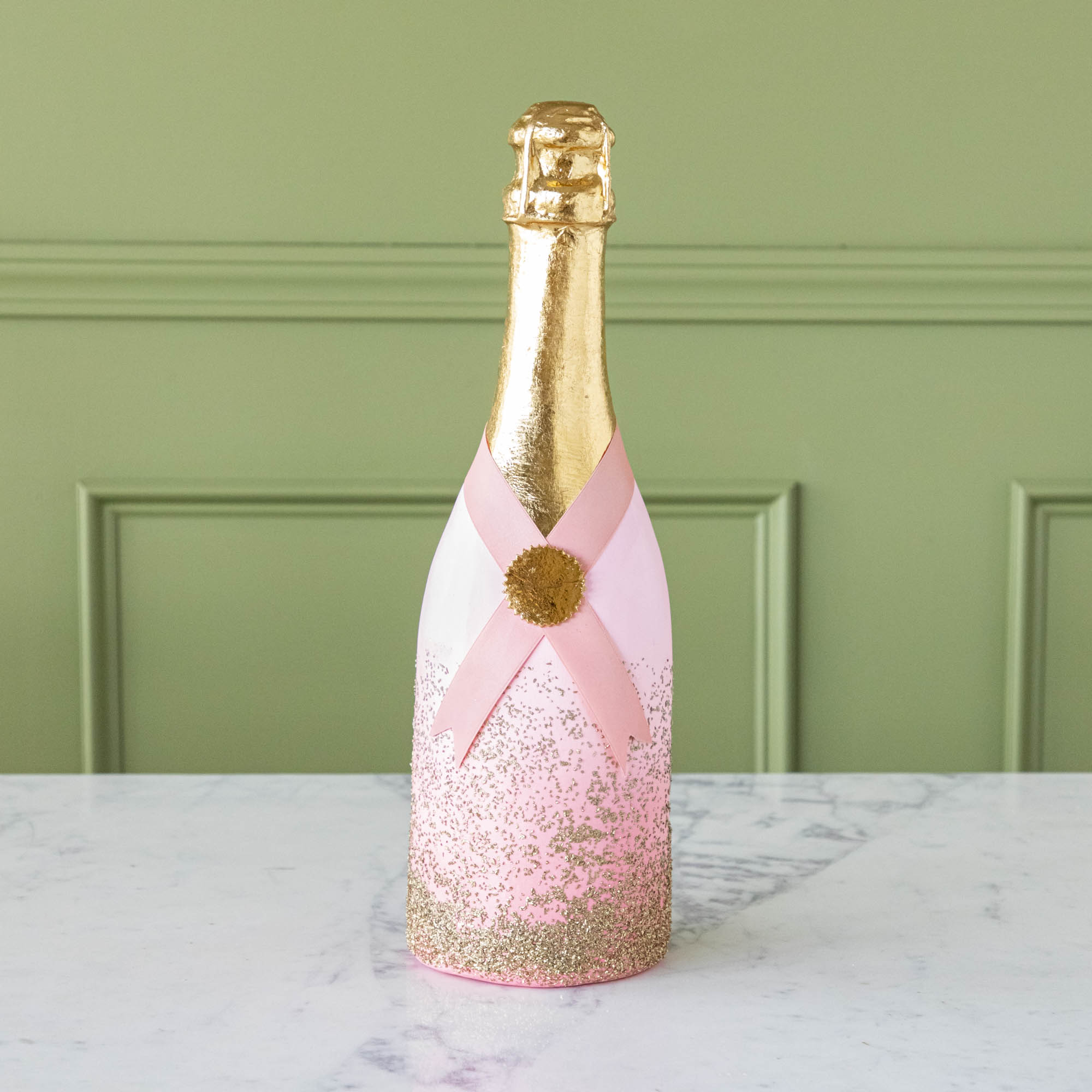 A pink, papermache Oversized Champagne bottle with a gold leafed top, a pink ribbon wrapped around the bottle held together by a gold foil seal, and gold glitter going up the bottle.