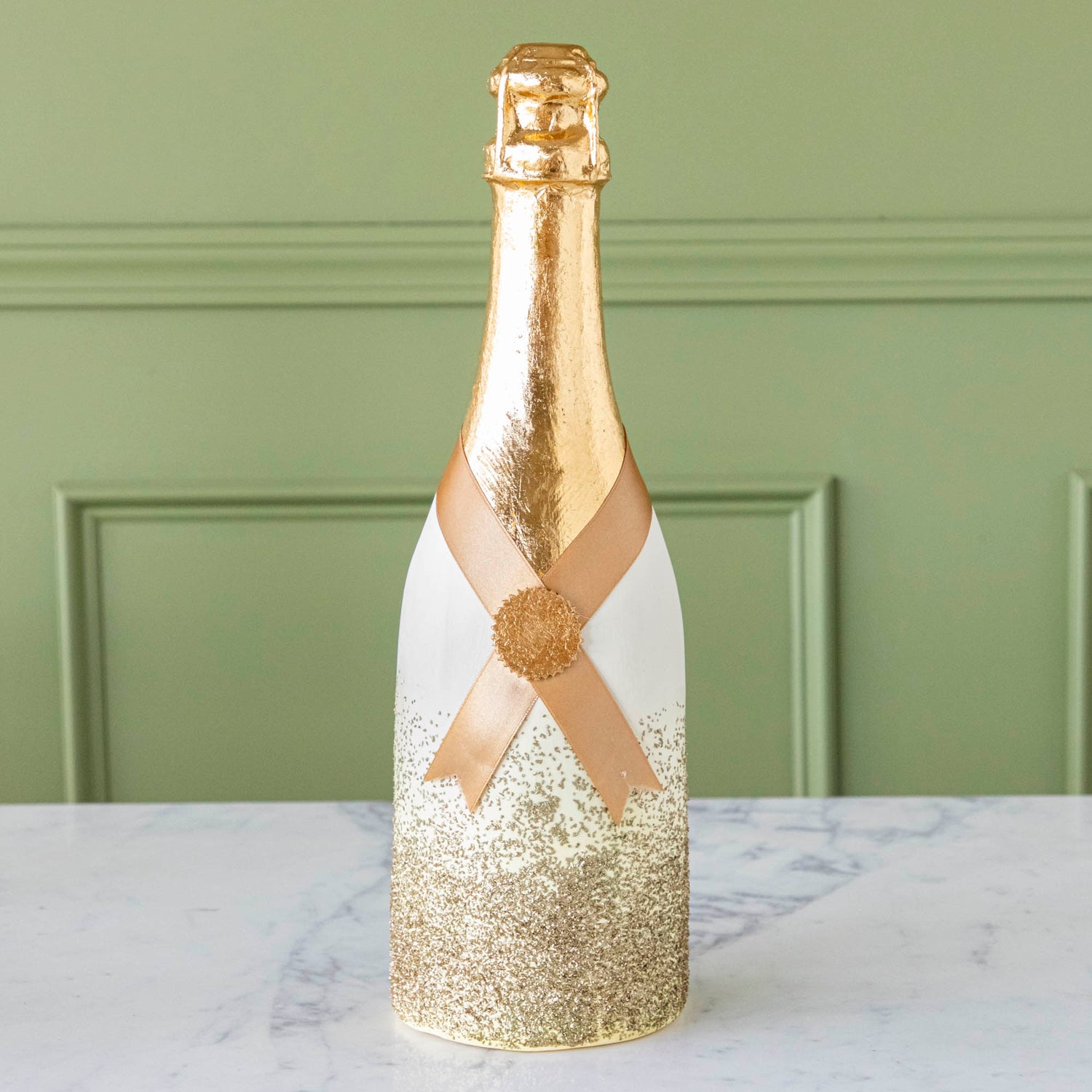 A gold, papermache Oversized Champagne bottle with a gold leafed top, a gold ribbon wrapped around the bottle held together by a gold foil seal, and gold glitter going up the bottle.