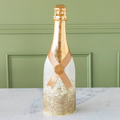 A gold, papermache Oversized Champagne bottle with a gold leafed top, a gold ribbon wrapped around the bottle held together by a gold foil seal, and gold glitter going up the bottle.