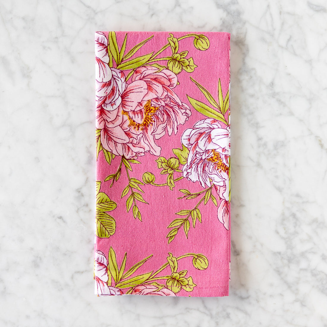 A pink cotton napkin, made in India, features a colorful illustration of pink flowers and green leaves and is displayed on a marble table.