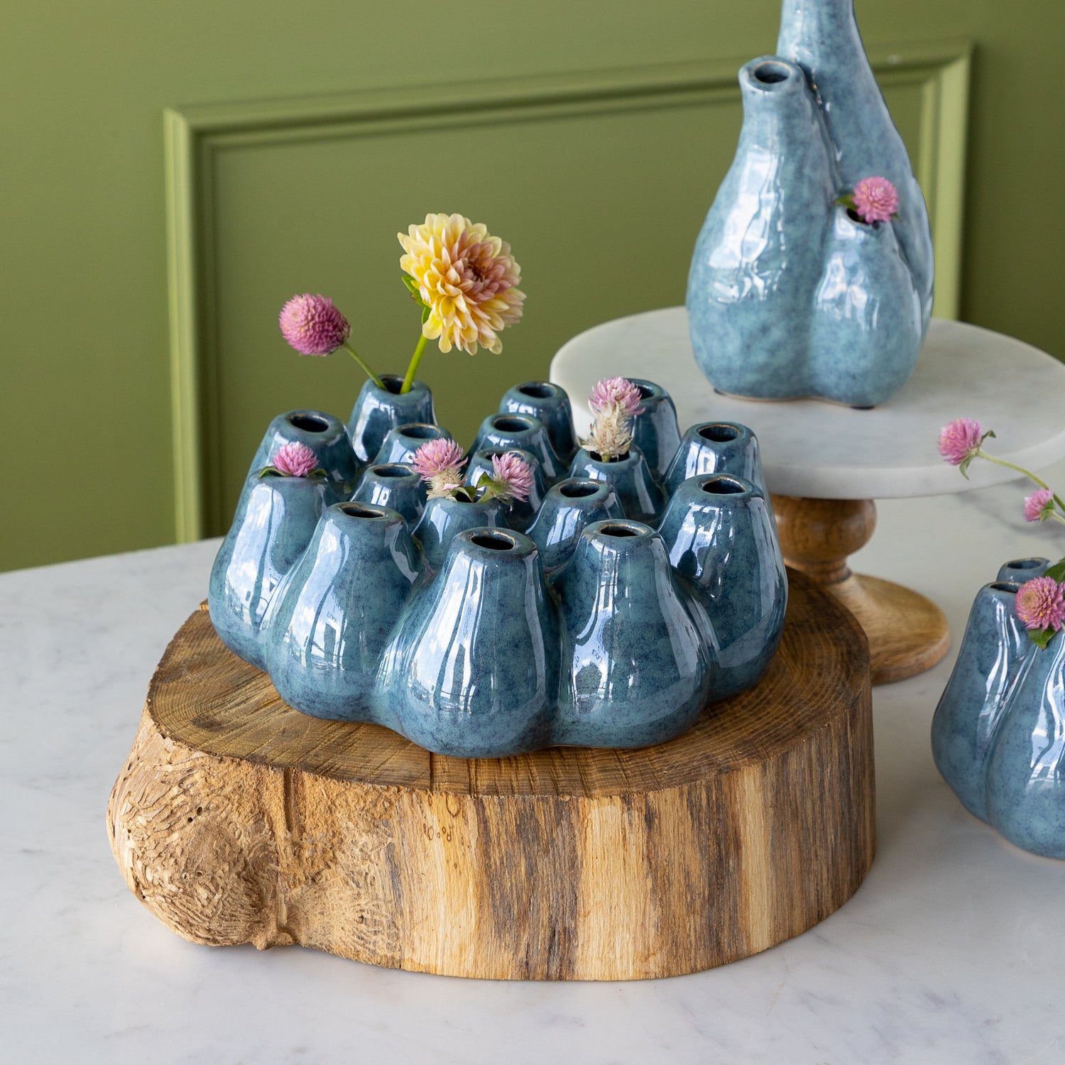A blue ceramic vase with flowers on a wooden stand against a green wall.
