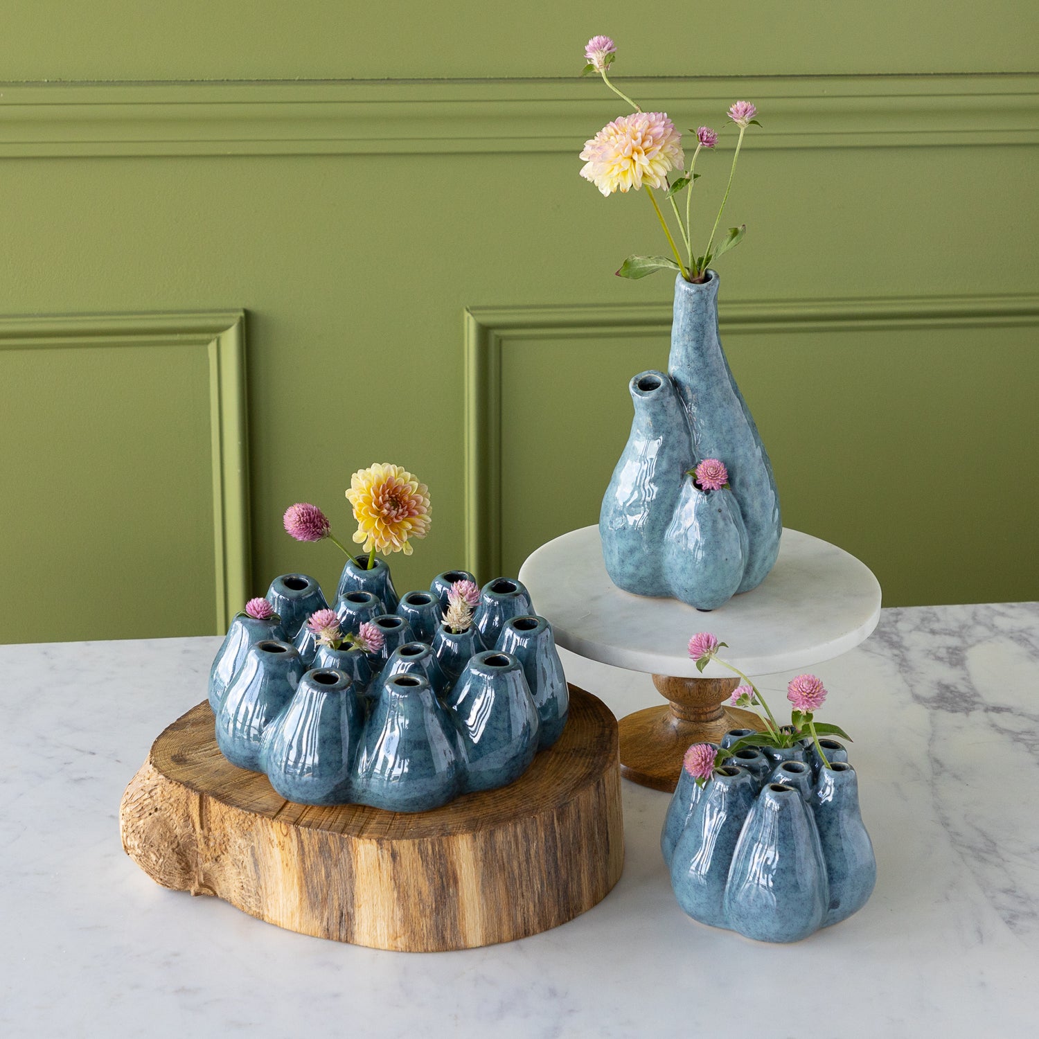 Set of blue ceramic vases with flowers on a marble surface against a green wall.