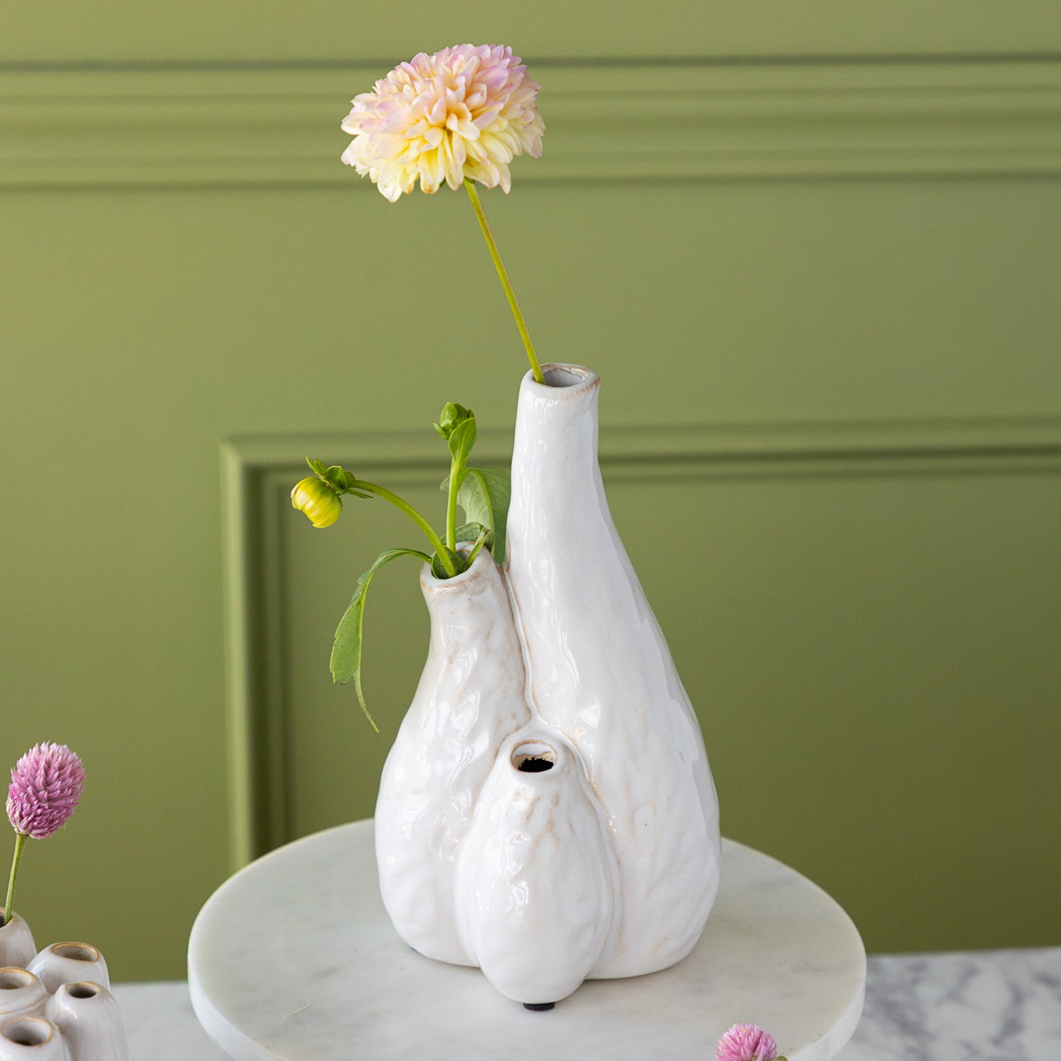 A white ceramic vase with three holes and flowers against a green wall.