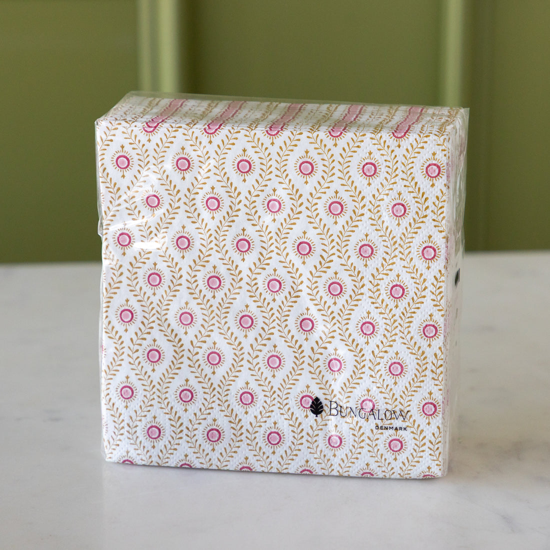 A square pack of Bungalow paper napkins featuring a white background with a repeating pattern of pink circles and gold leafy motifs, wrapped in clear plastic on a white marble surface with a green background.