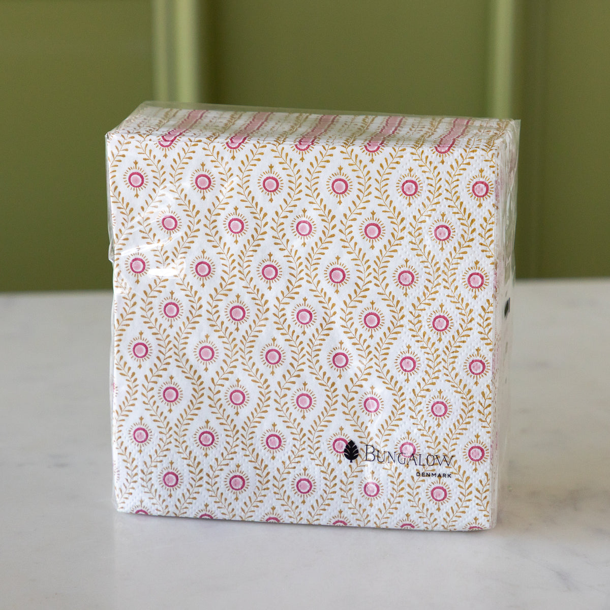 A square pack of Bungalow paper napkins featuring a white background with a repeating pattern of pink circles and gold leafy motifs, wrapped in clear plastic on a white marble surface with a green background.