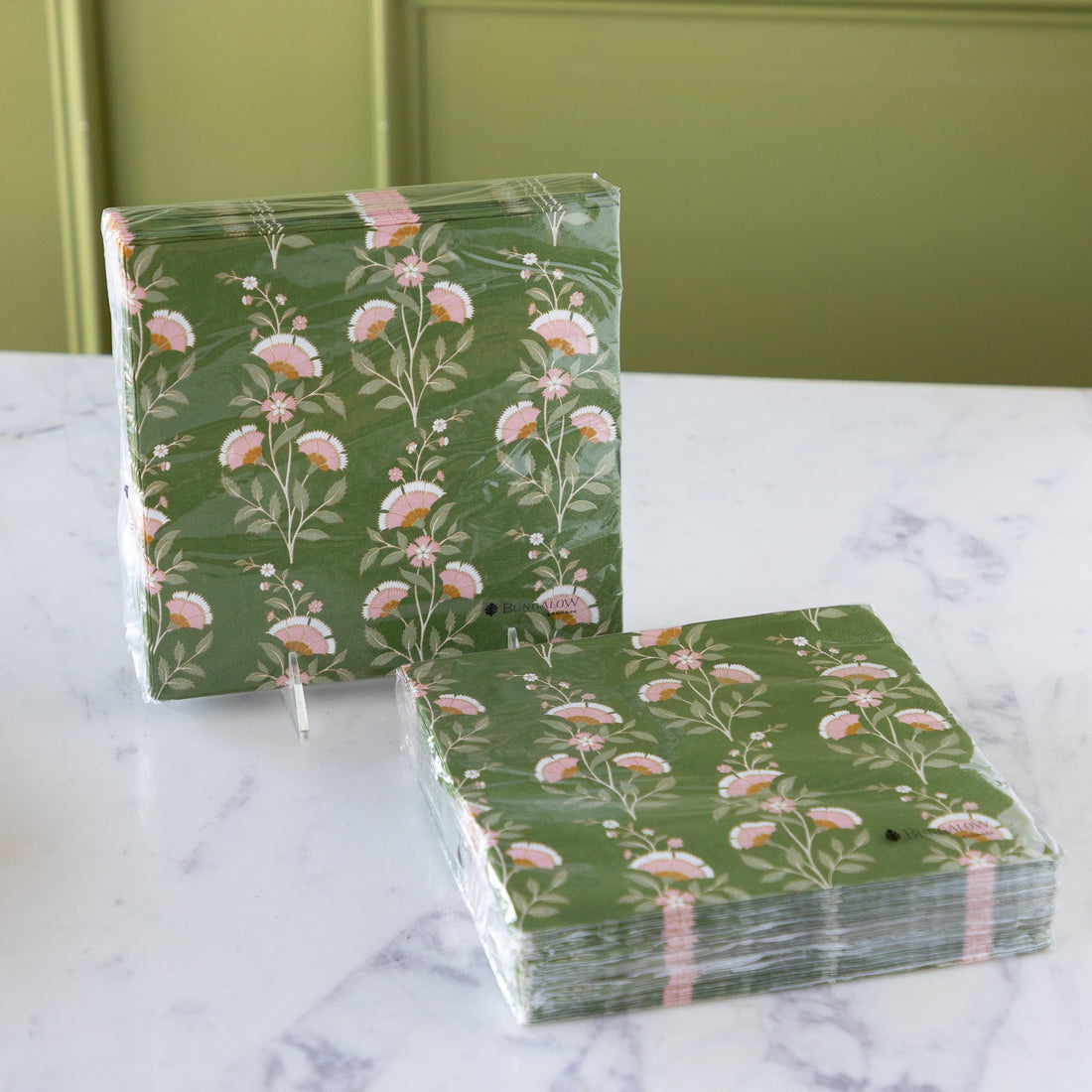 Two stacks of green floral paper napkins wrapped in clear plastic, displayed on a white marble surface with a green paneled background.