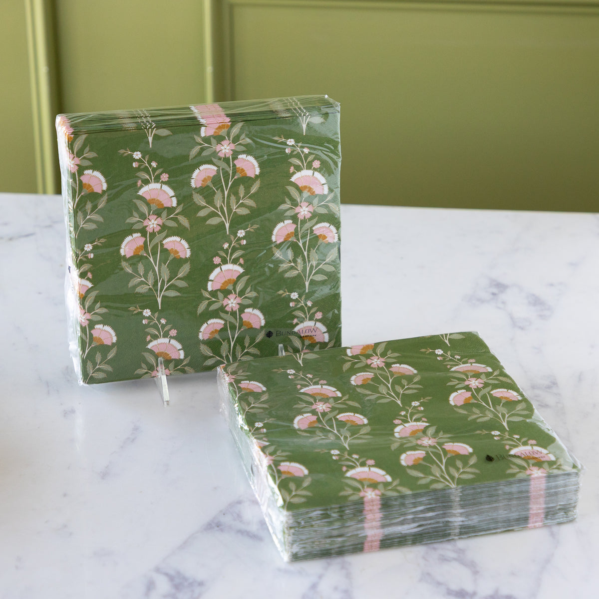 Two stacks of green floral paper napkins wrapped in clear plastic, displayed on a white marble surface with a green paneled background.