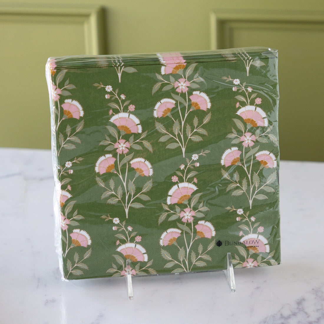 A stack of square paper napkins wrapped in clear plastic, featuring a green background with a pink and white floral pattern, displayed on a marble surface against a green paneled wall.