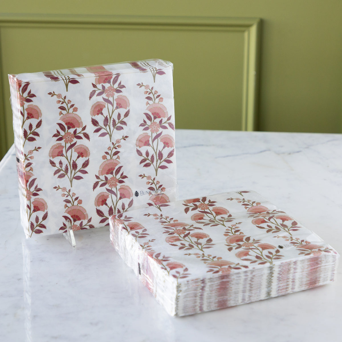 A stack of rectangular paper napkins with a pink and burgundy floral vine pattern on a white background, displayed on a white marble tabletop against an olive green paneled wall.
