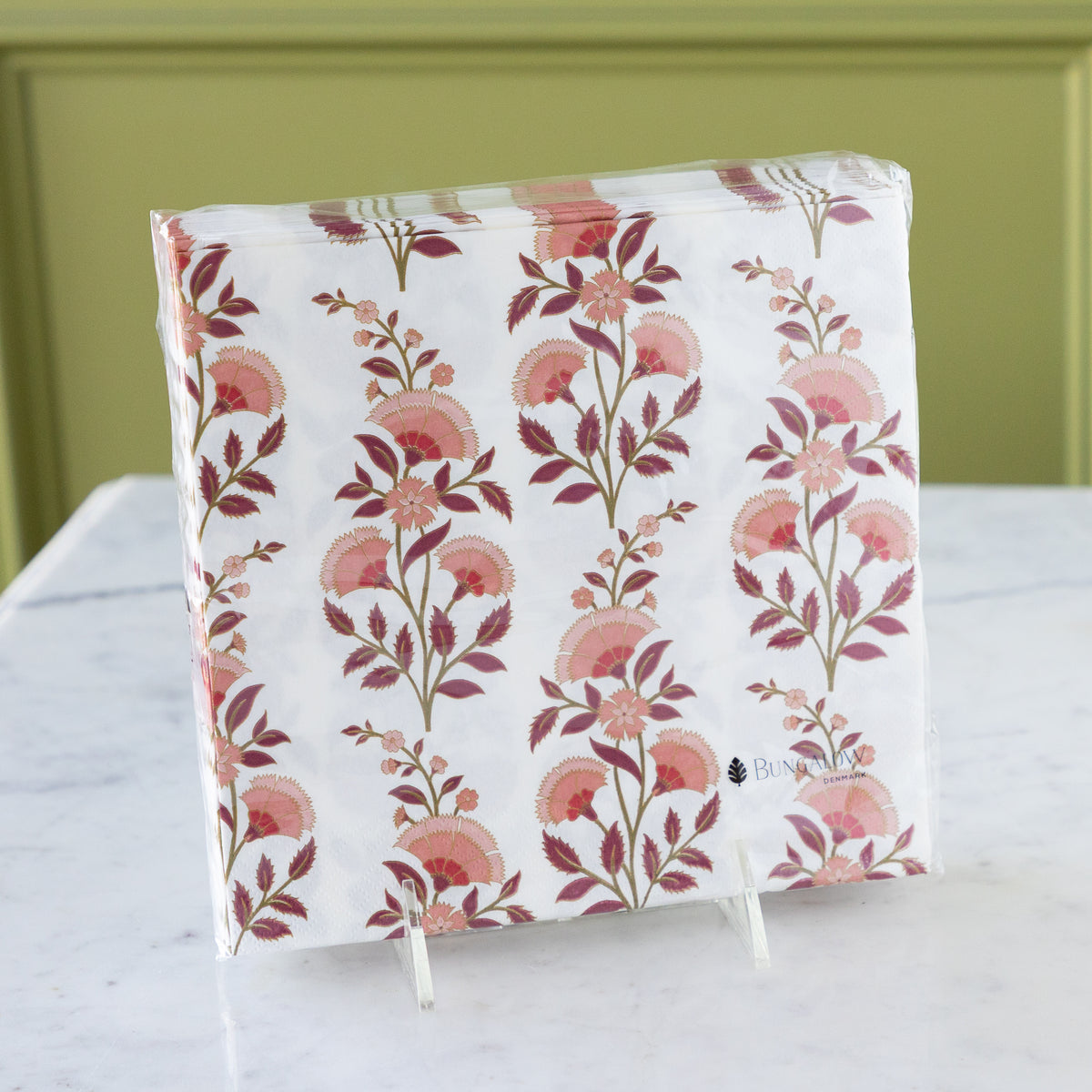 A packaged set of white napkins with a vertical floral pattern in dusty pink and burgundy, displayed on a small white stand on a white marble surface against a green paneled background.