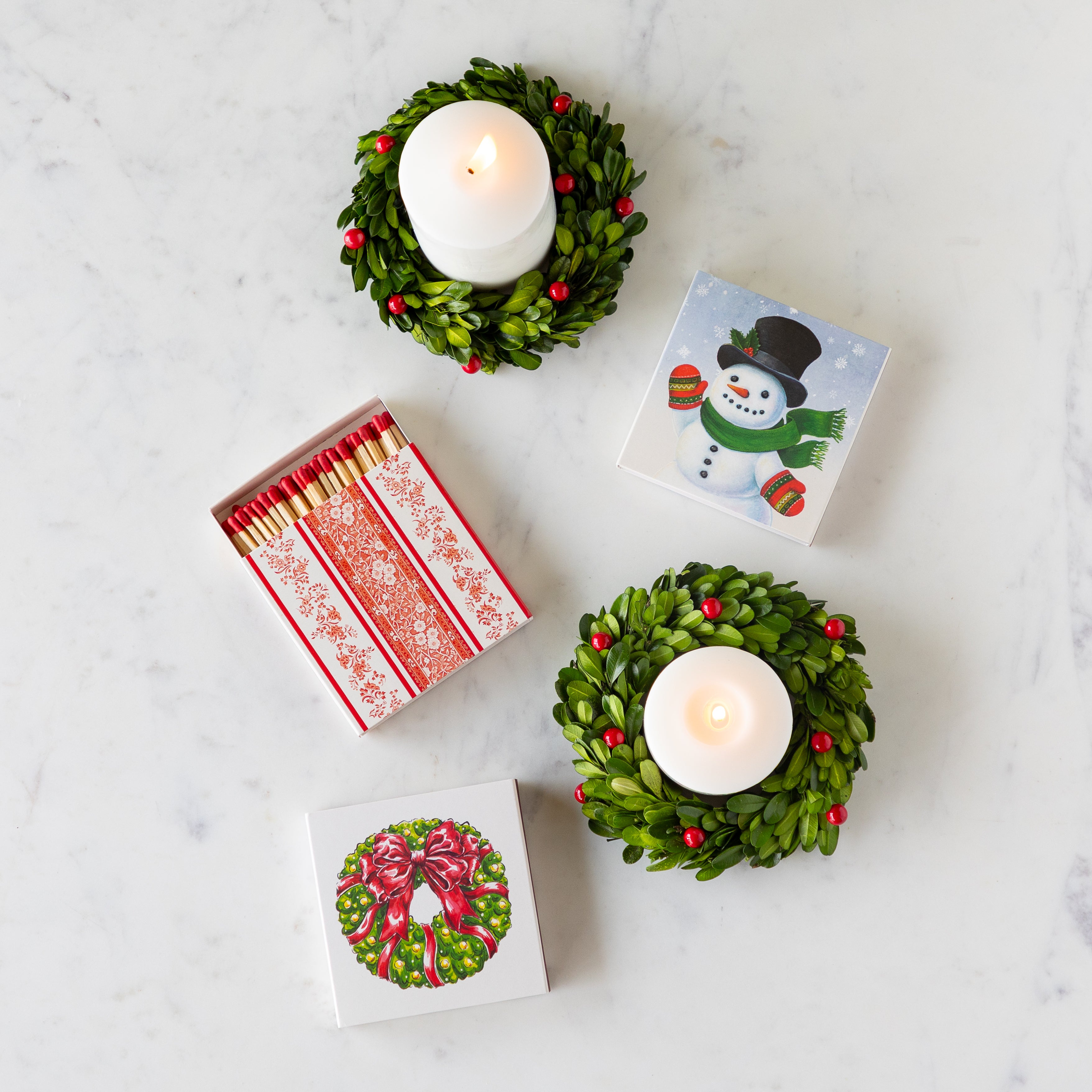 The Red Burleigh, Snowman and Holiday Wreath Matches arranged on a table next to lit pillar candles with a boxwood wreath with berries around them.