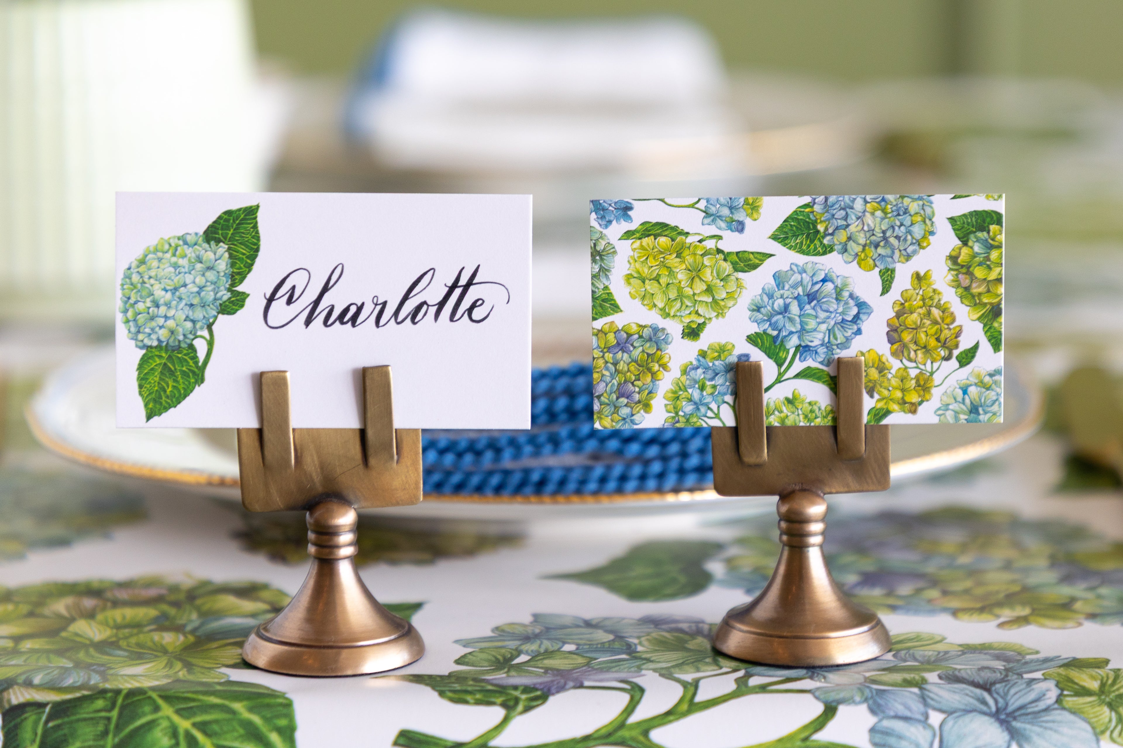 Two decorative place cards with floral hydrangea prints are displayed on small brass stand holders on a patterned tablecloth in a styled dining setting.