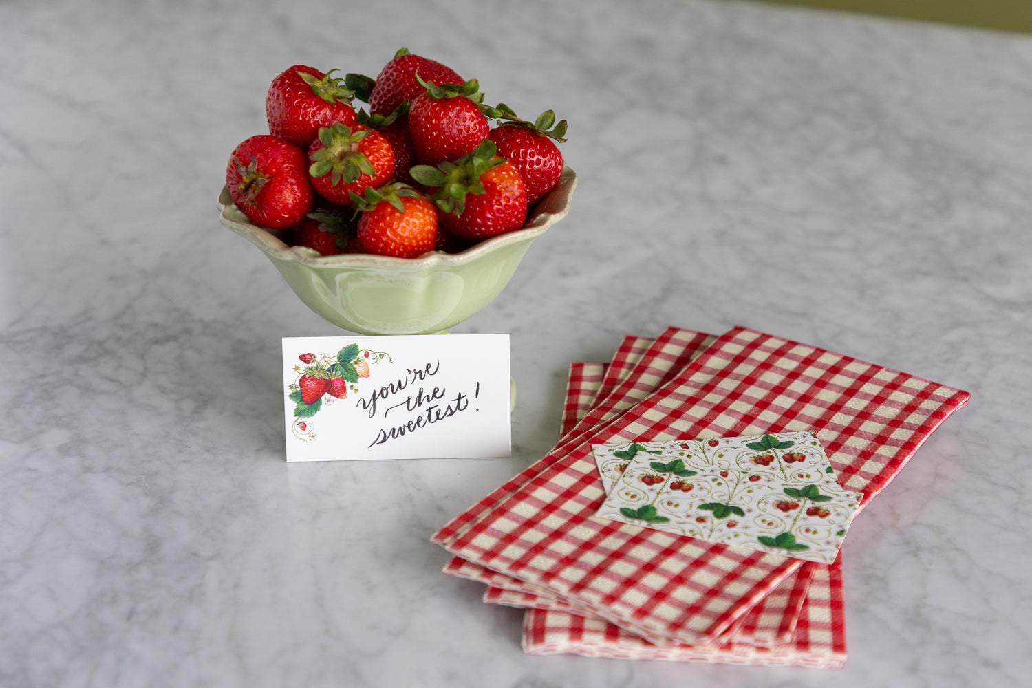 A styled tabletop arrangement featuring a bowl of fresh red strawberries, a small handwritten note that reads &