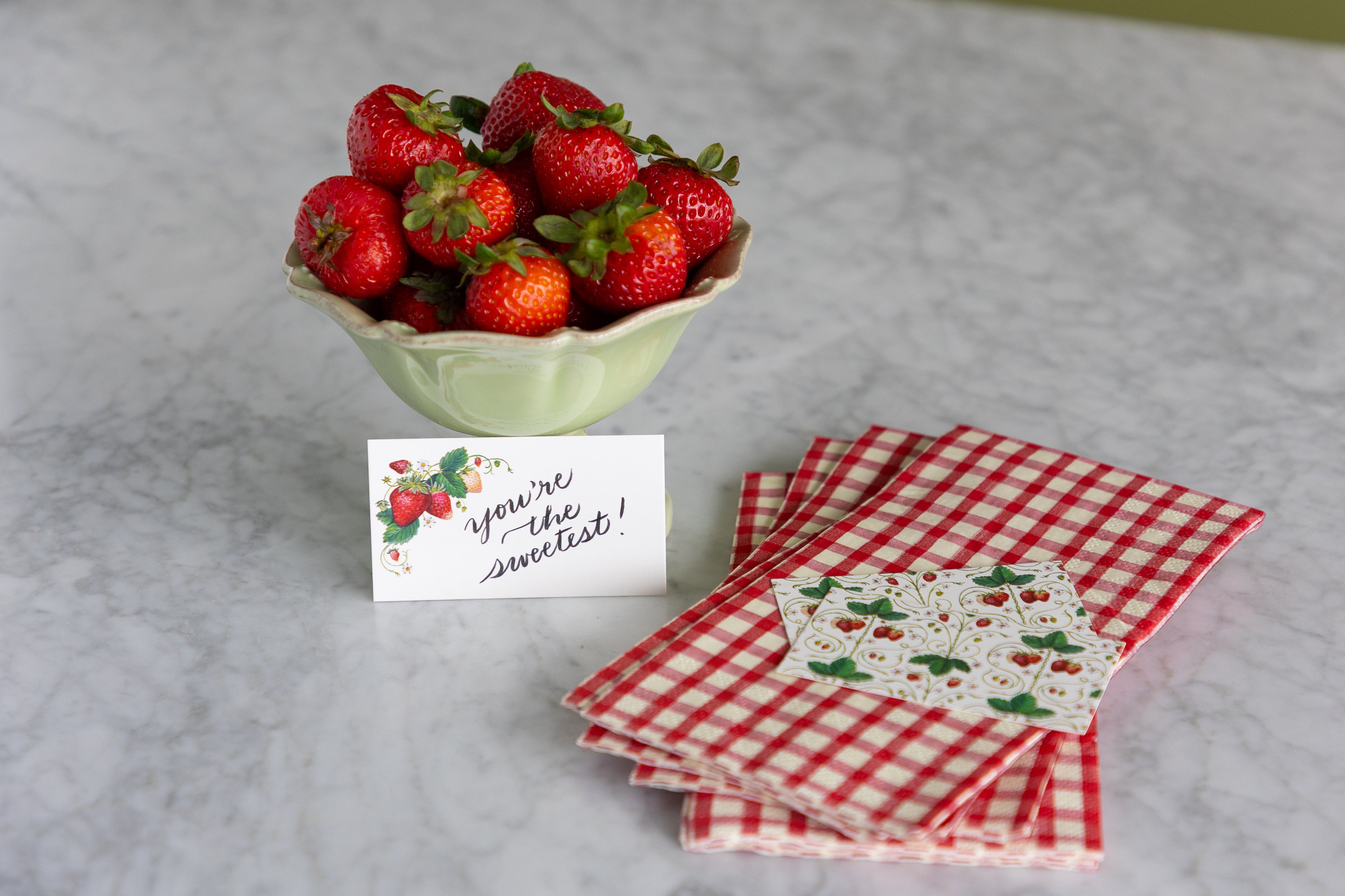 A styled tabletop arrangement featuring a bowl of fresh red strawberries, a small handwritten note that reads &