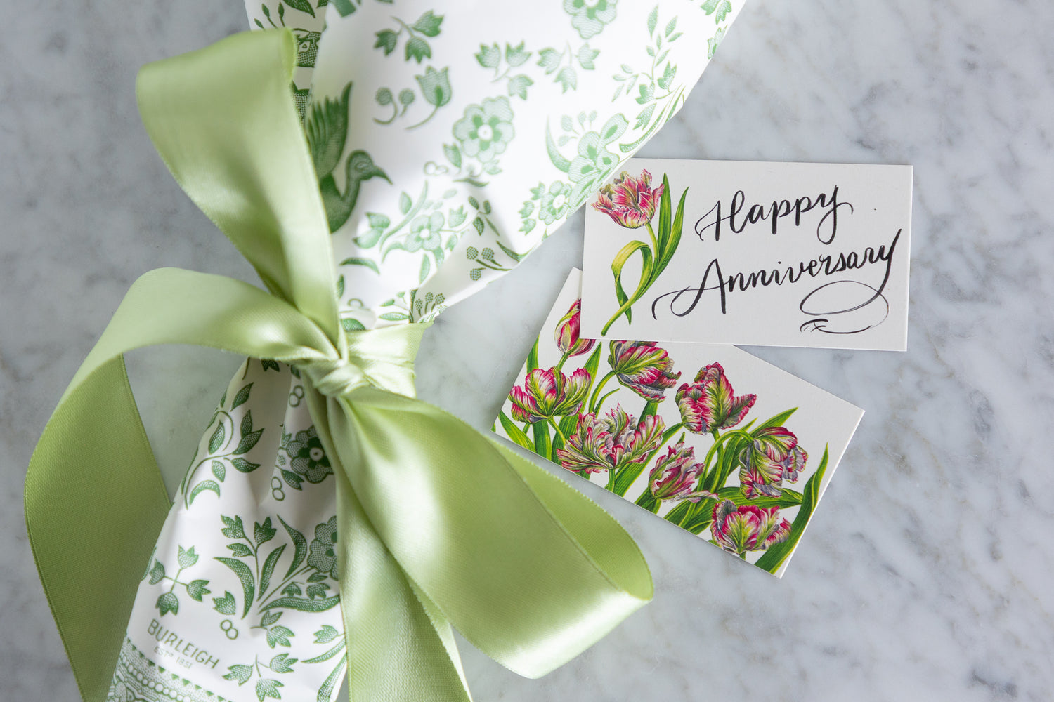 A wrapped gift tied with a pale green satin ribbon, featuring white wrapping paper with a green botanical pattern and a small matching floral card that reads &
