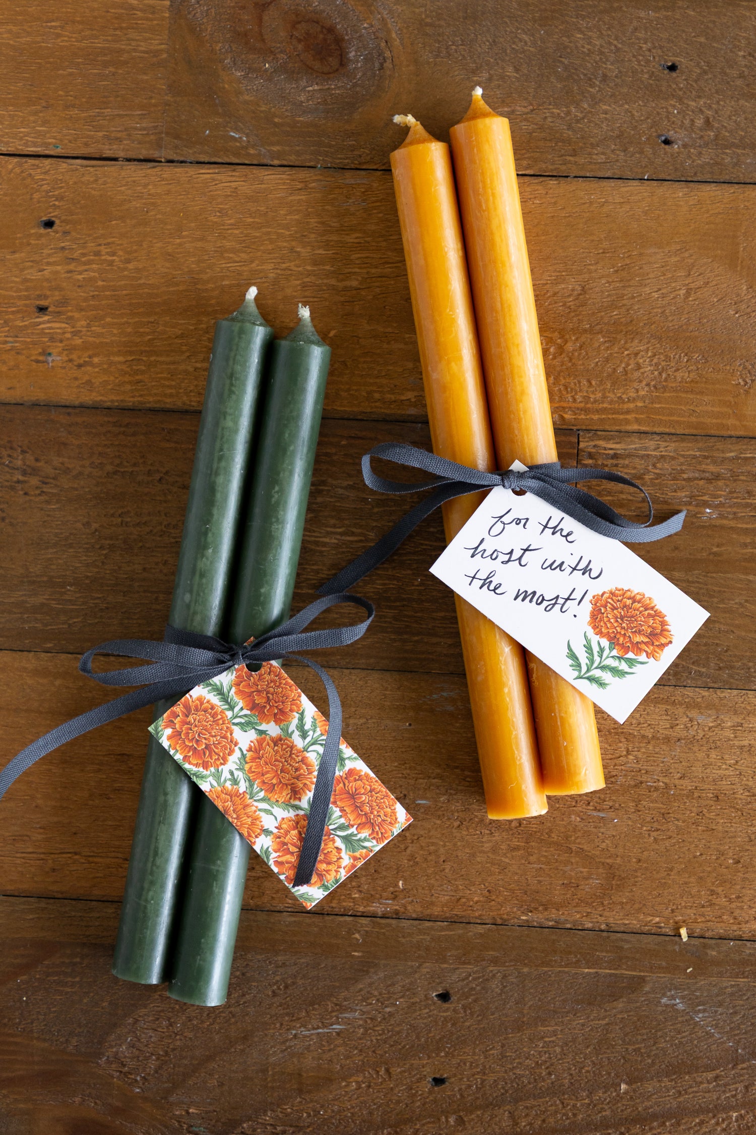 Two bundles of taper candles on a rustic wooden surface. One bundle contains dark green candles tied with a dark ribbon and a floral tag, and the other contains golden yellow candles tied with a navy ribbon and a handwritten tag that reads &