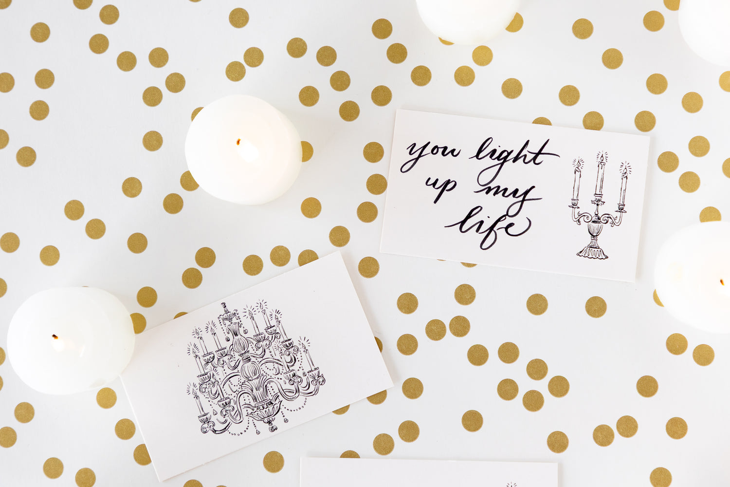 Two white rectangular cards with black printed illustrations and text, arranged on a white surface with scattered gold polka dots and white candles.