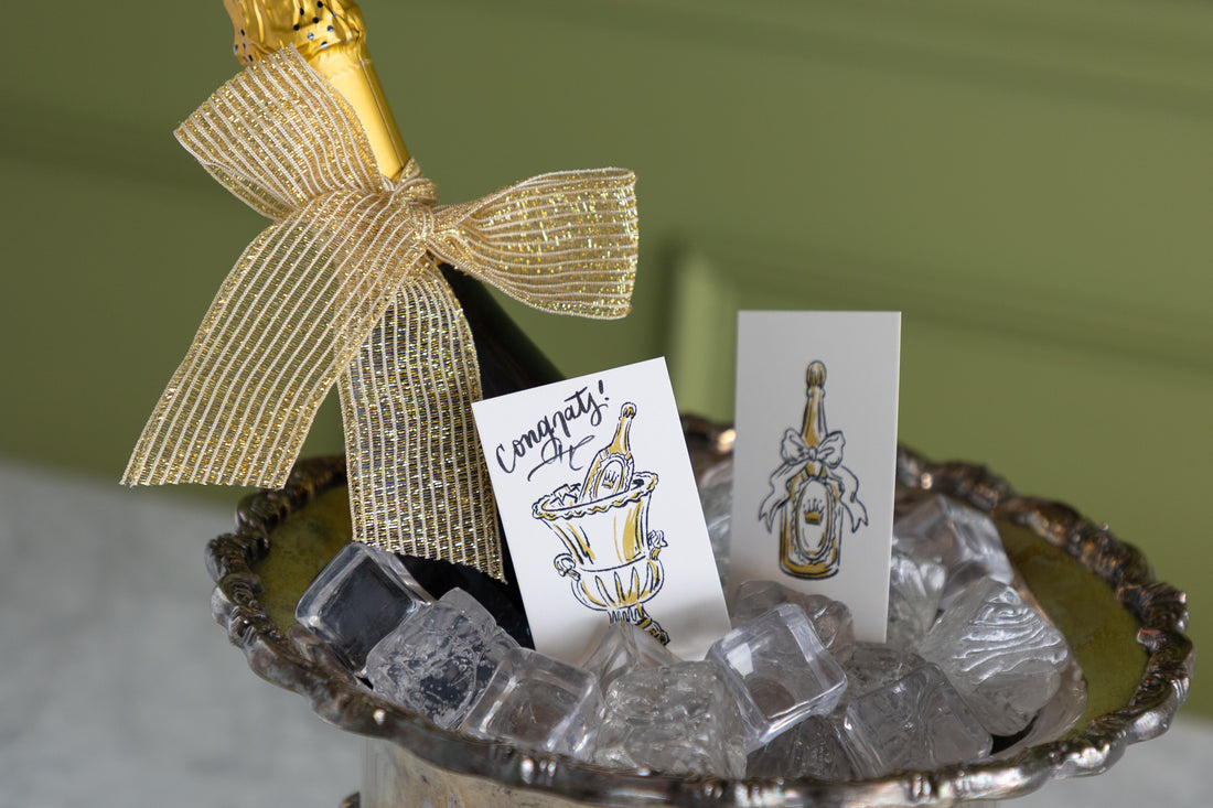 A champagne bottle with a gold foil top and a large gold ribbon bow sits in a silver tray filled with ice cubes, alongside two printed cards featuring illustrated champagne bottles and one card that says &