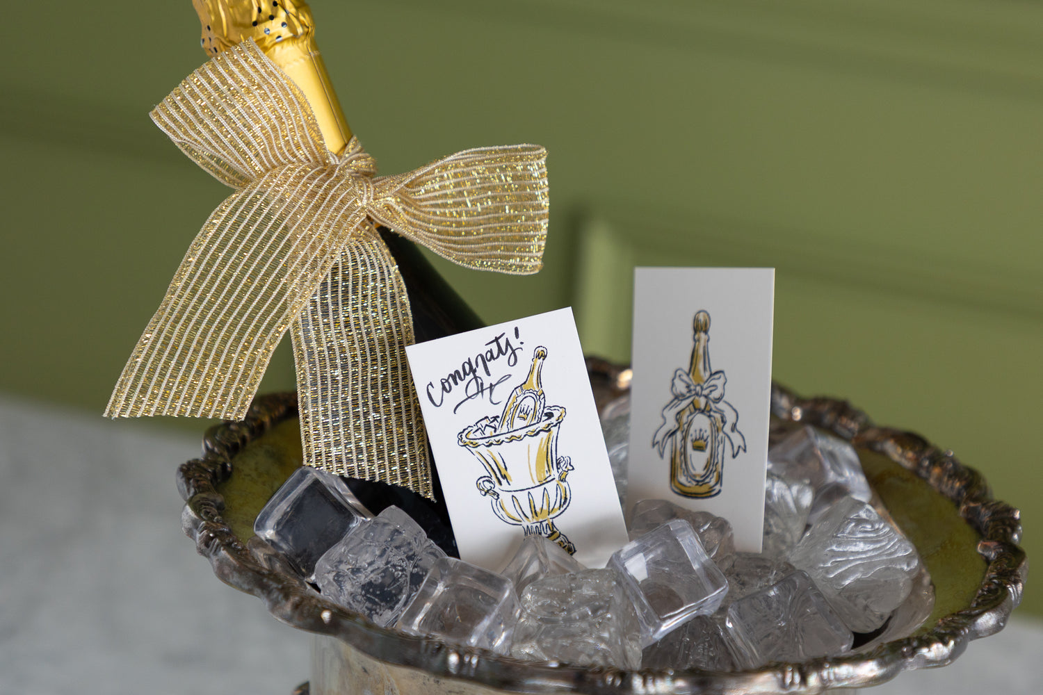 A champagne bottle with a gold foil top and a large gold ribbon bow sits in a silver tray filled with ice cubes, alongside two printed cards featuring illustrated champagne bottles and one card that says &
