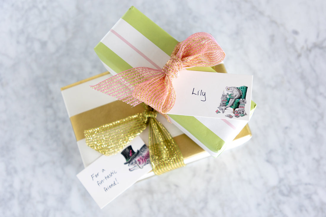A styled gift box wrapped in light green and white striped paper, gold foil, and a peach mesh bow, with two handwritten gift tags on a light gray marble surface.
