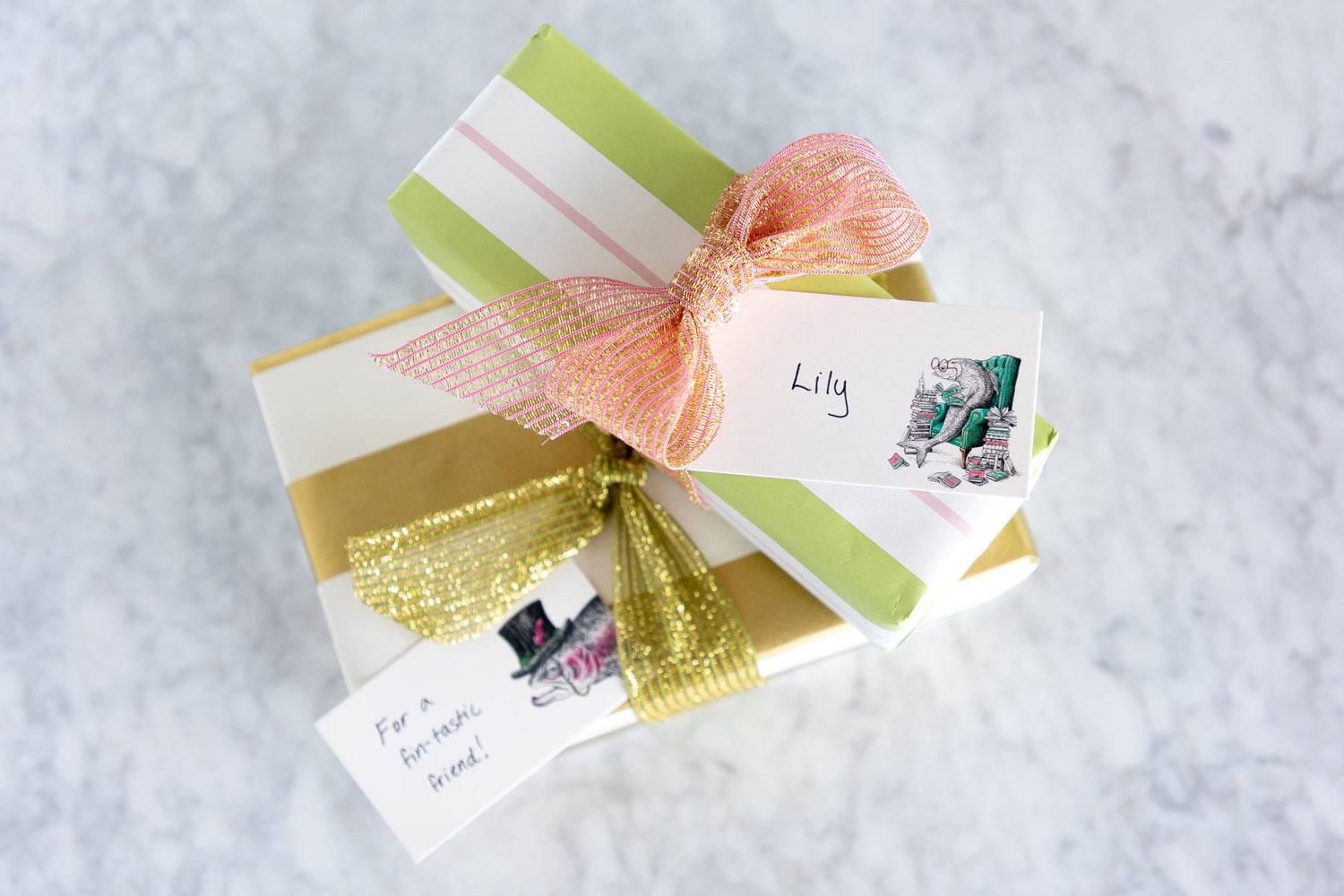 A styled gift box wrapped in light green and white striped paper, gold foil, and a peach mesh bow, with two handwritten gift tags on a light gray marble surface.