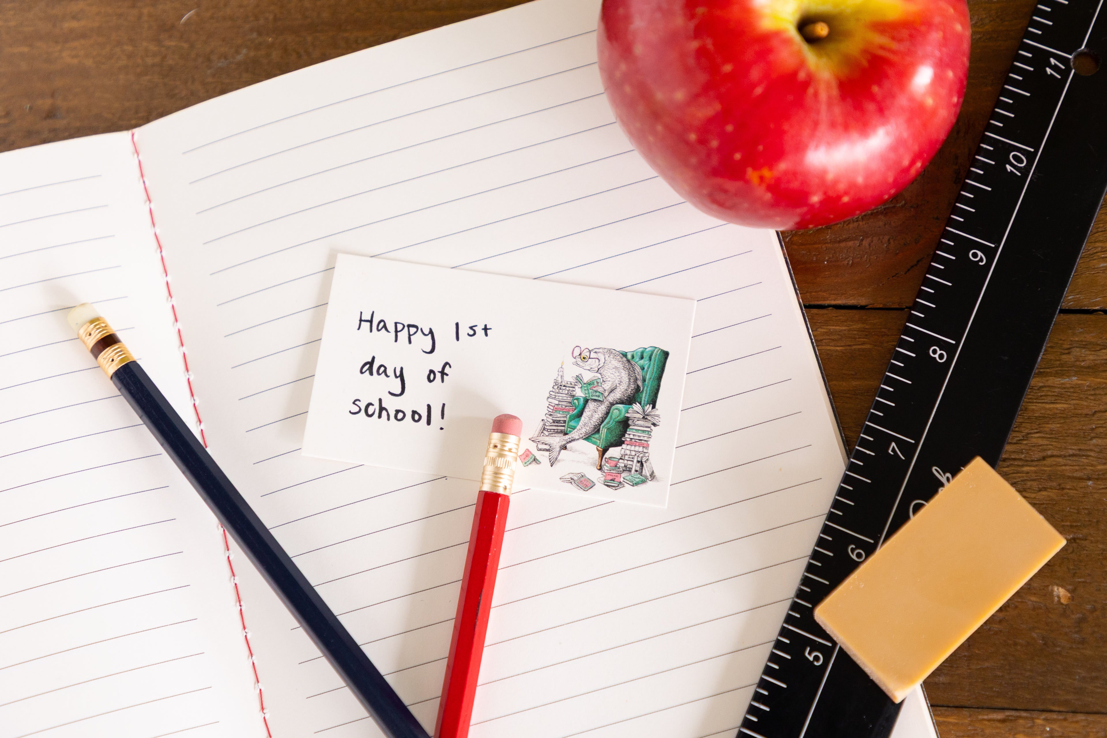 A styled flat-lay of school supplies on a wooden table, including an open lined notebook, a handwritten &