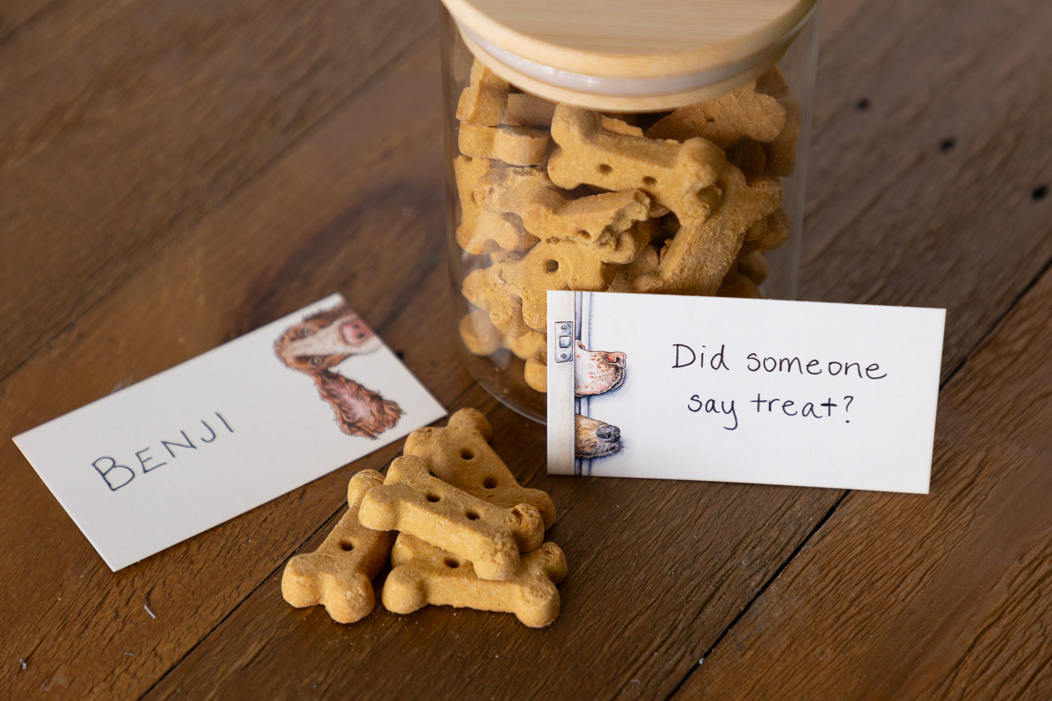 A clear glass jar filled with bone-shaped dog treats sits on a wooden table, with a few treats placed in front and two handwritten cards nearby reading &
