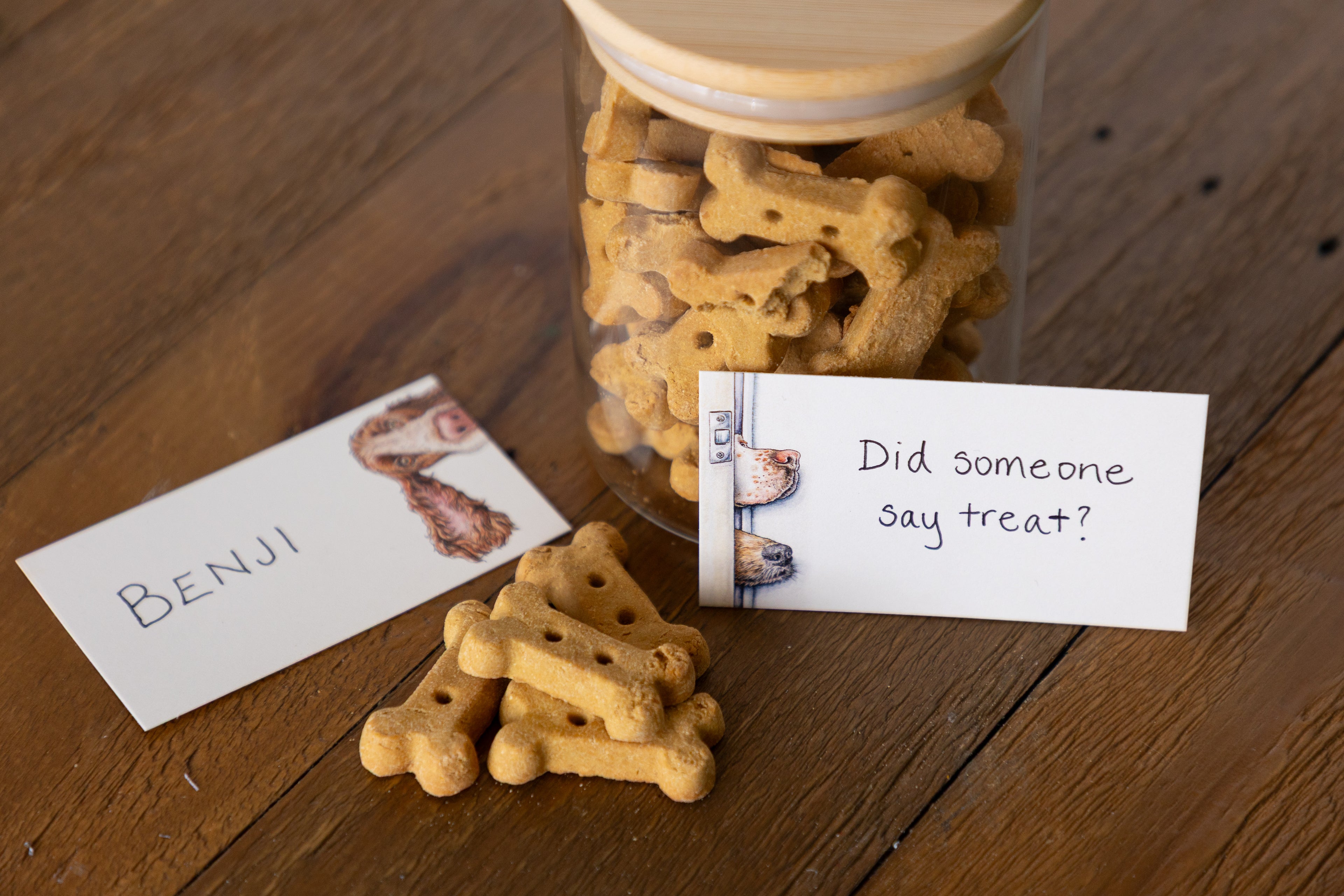 A clear glass jar filled with bone-shaped dog treats sits on a wooden table, with a few treats placed in front and two handwritten cards nearby reading &