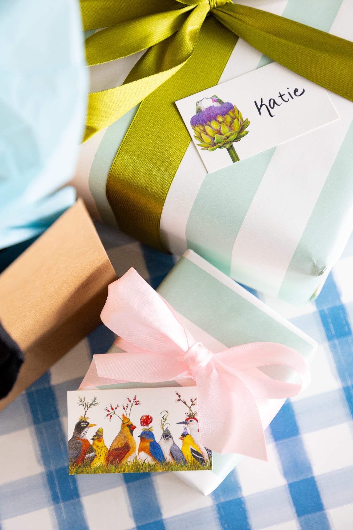 A close-up of wrapped gift boxes on a blue-and-white checkered surface. One box has white and mint green stripes with a large olive-green ribbon and a name tag reading Katie with a purple artichoke illustration.
